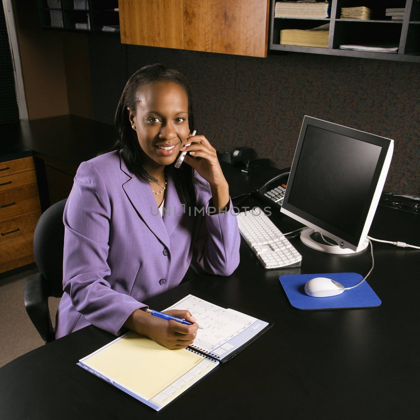 African-American young adult business woman talking on cell phone and writing in planner in office smiling and looking at viewer.