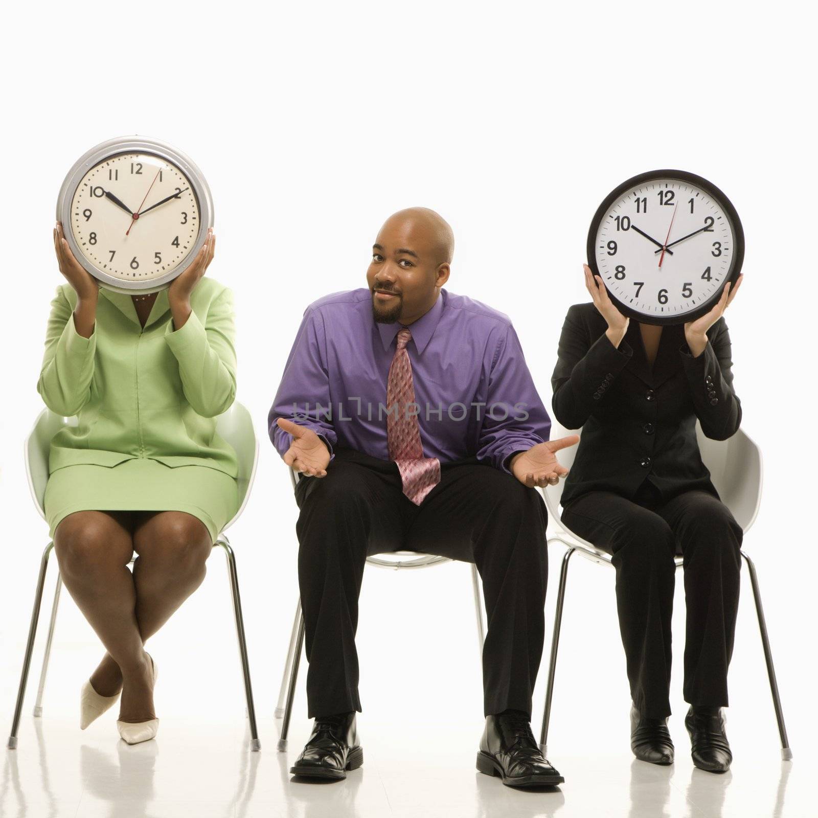 Businesswomen sitting holding clocks over faces while African-American businessman shrugs.