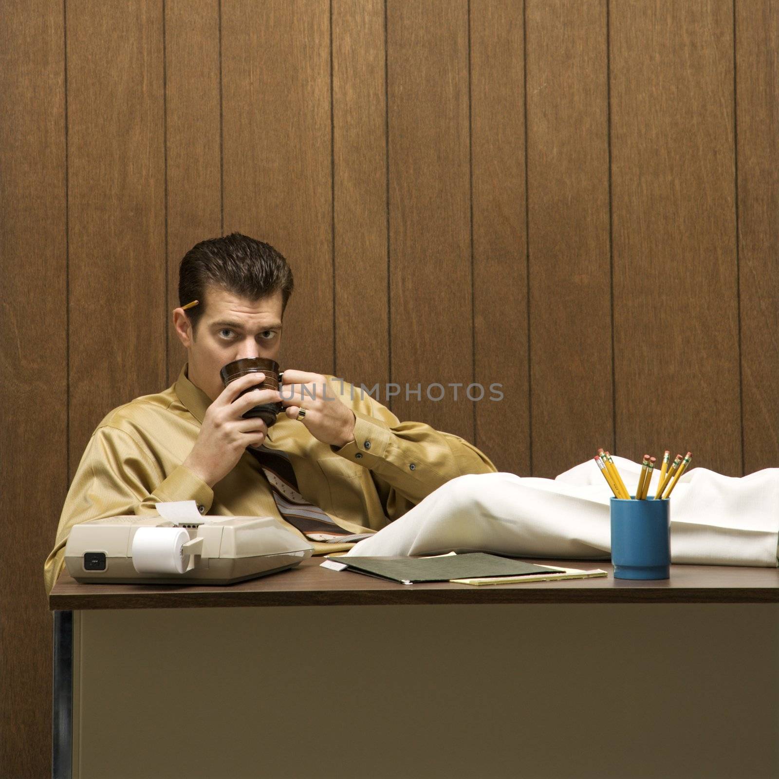 Caucasion mid-adult retro businessman sitting with feet propped on desk drinking coffee.