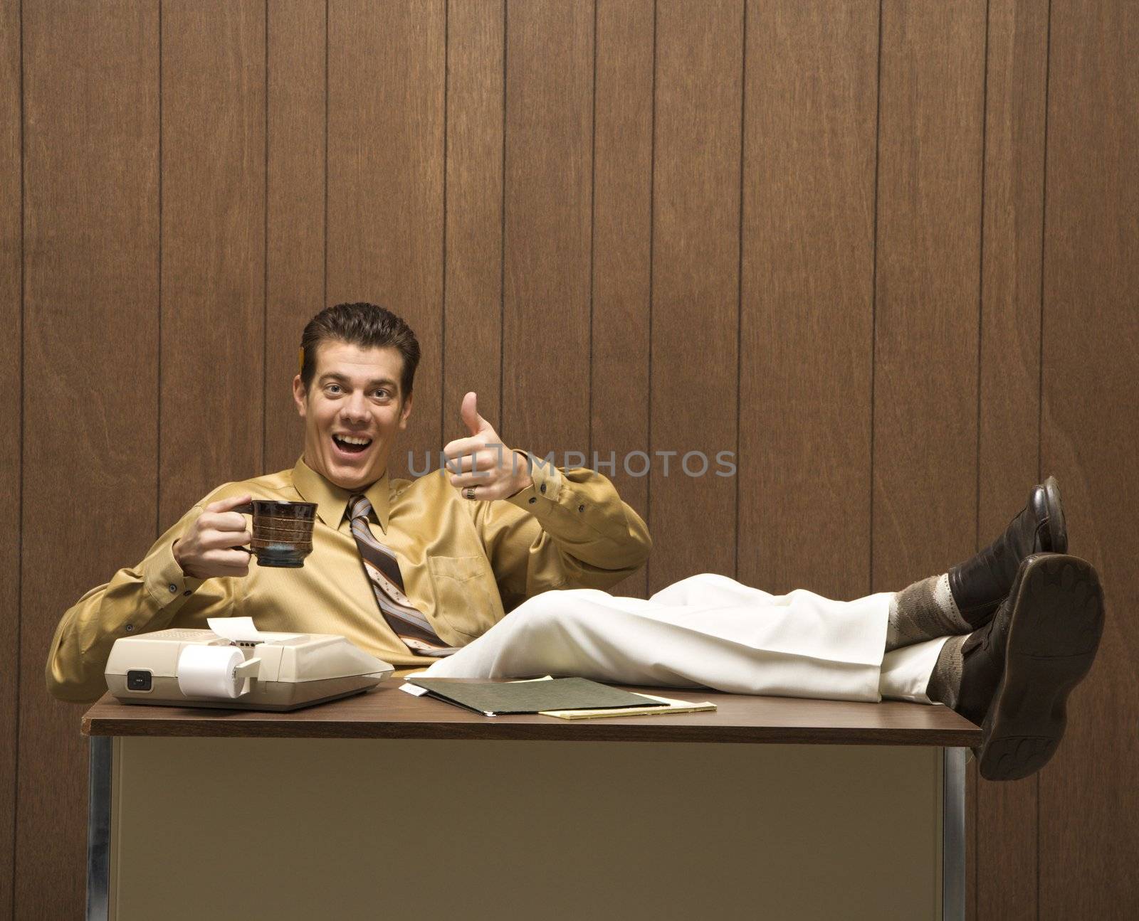Caucasion mid-adult retro businessman sitting with feet propped on desk drinking coffee giving a thumbs up.
