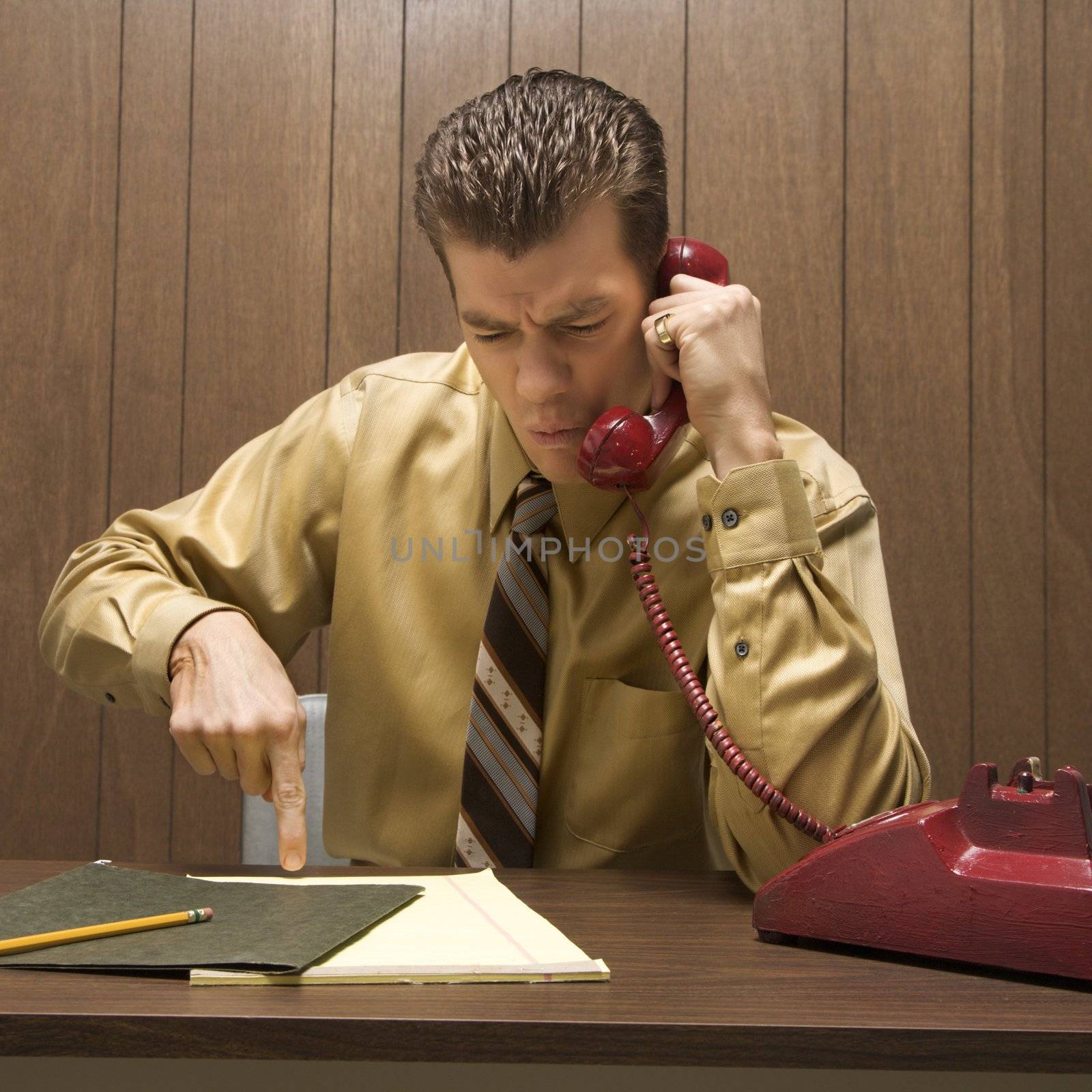 Caucasion mid-adult retro businessman sitting at desk talking on telephone with angry expression.