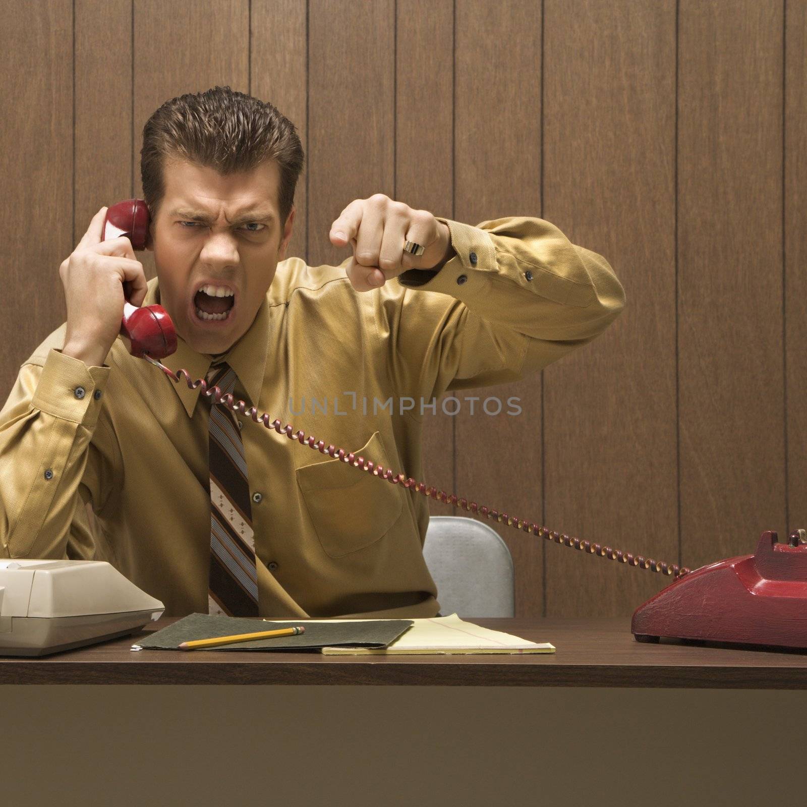 Caucasion mid-adult retro businessman sitting at desk talking on telephone with angry expression and gesture.