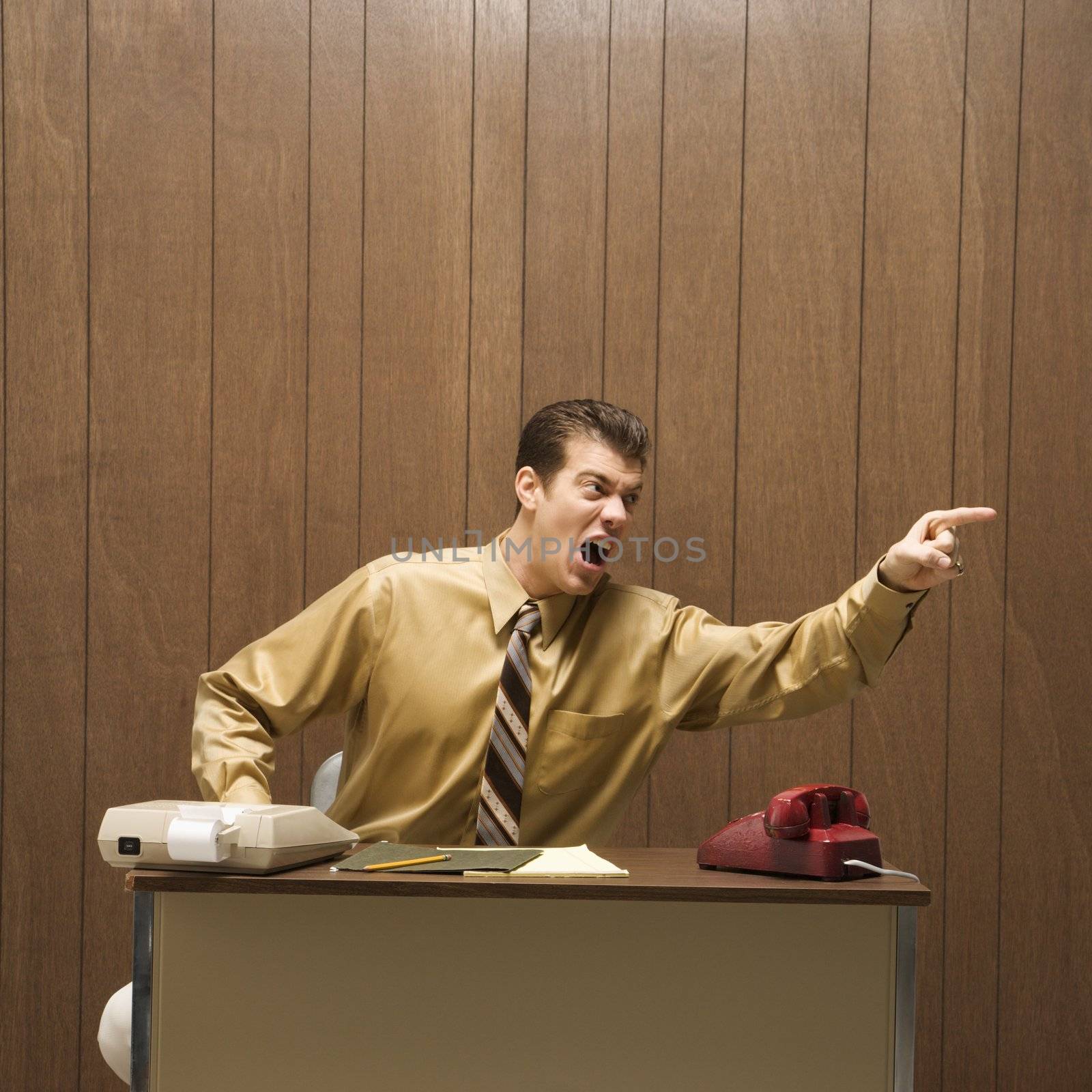 Caucasion mid-adult retro businessman sitting at desk pointing in anger.