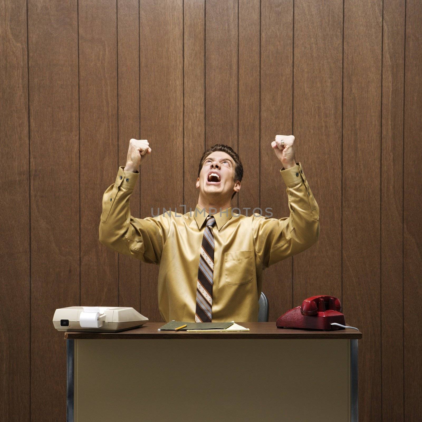 Caucasion mid-adult retro businessman sitting at desk raising fists in anger.