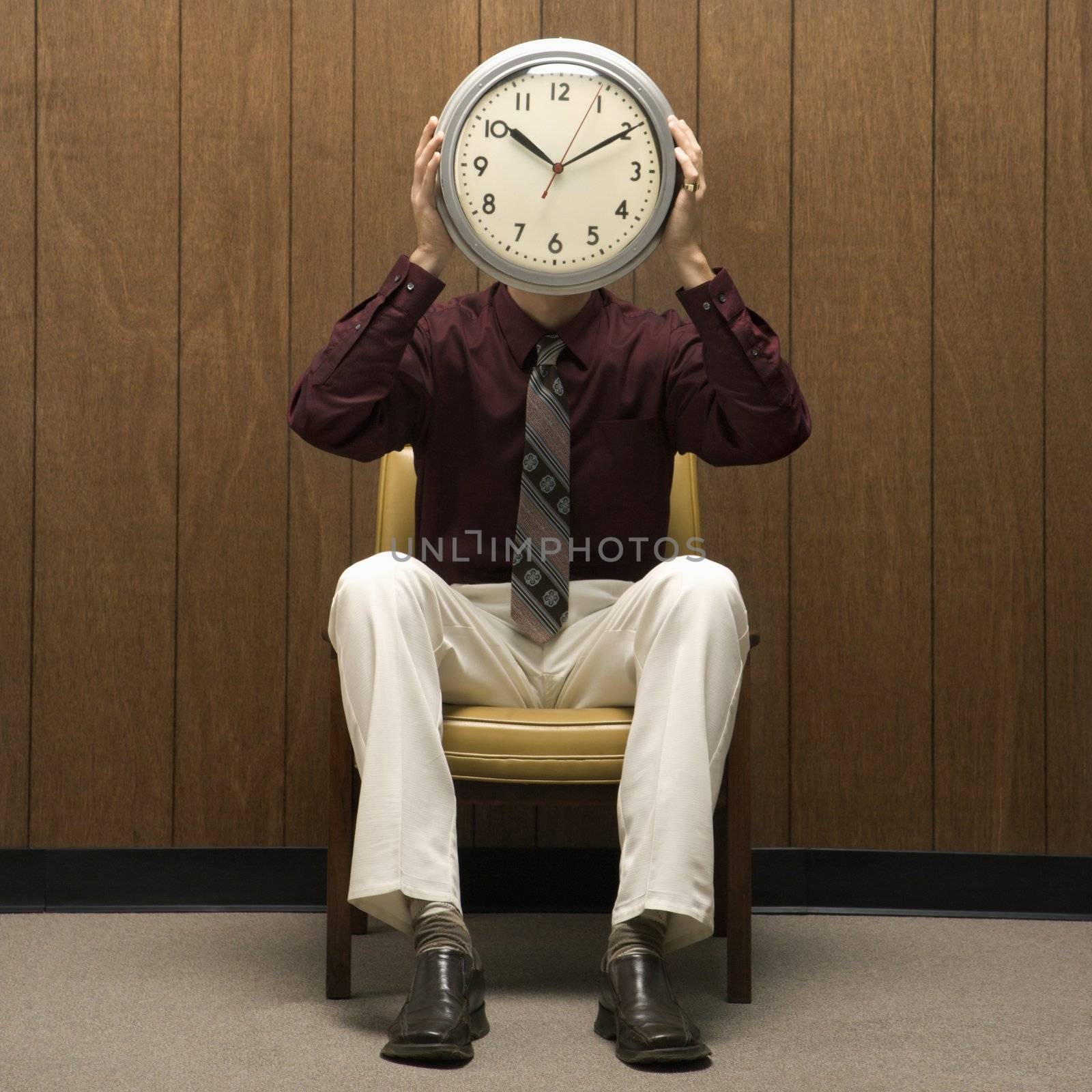 Caucasion mid-adult retro businessman sitting in chair against wood paneling holding clock over face.