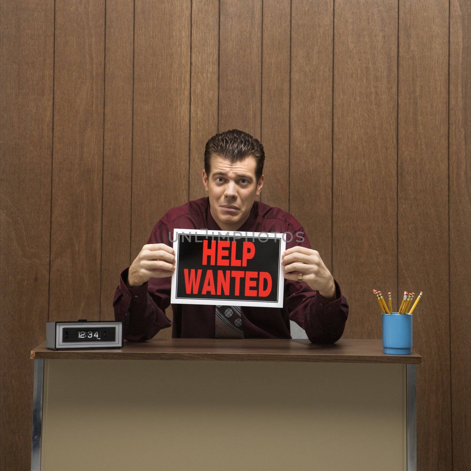 Caucasion mid-adult retro businessman with desperate expression sitting at desk holding up help wanted sign .