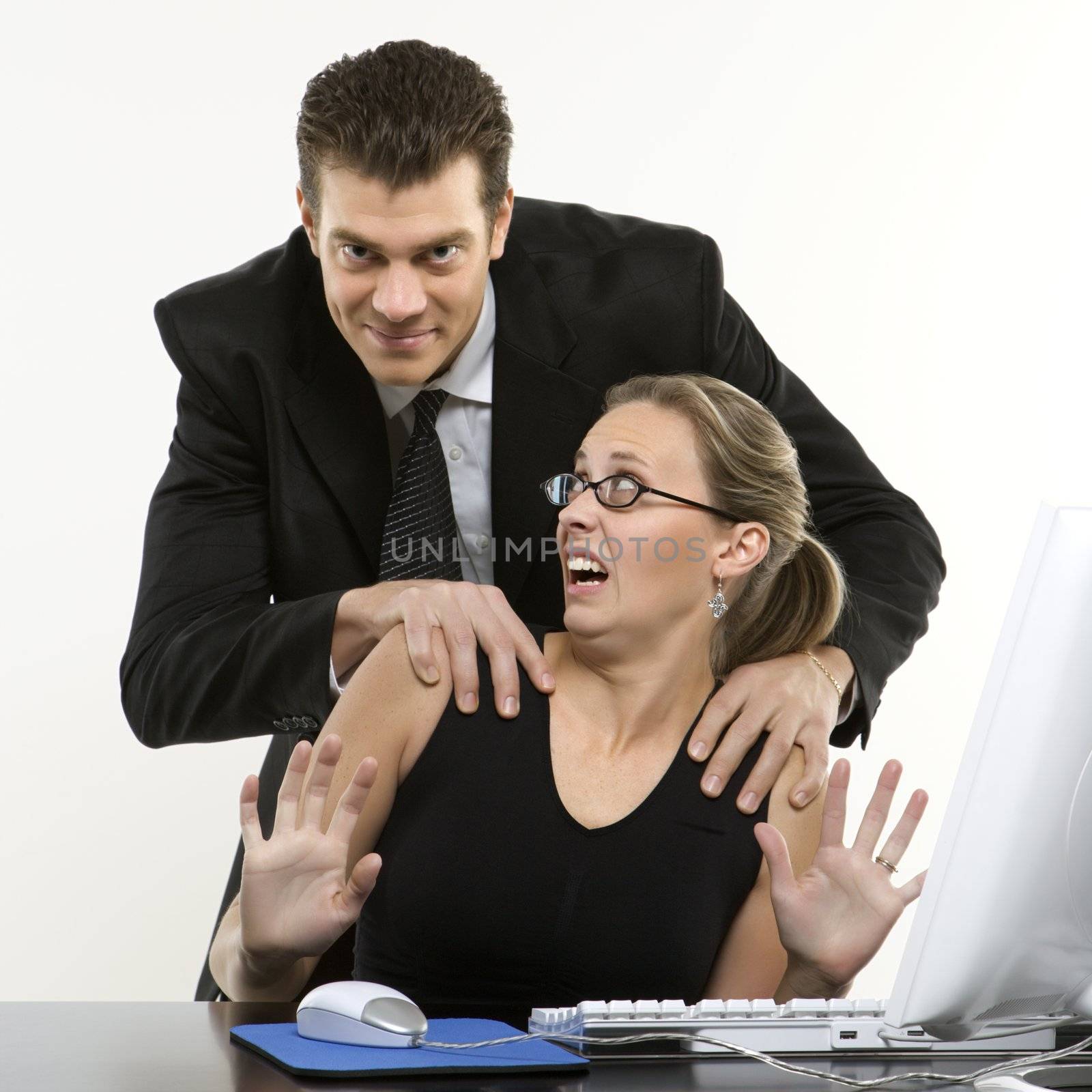 Caucasian mid-adult man sexually harassing woman sitting at computer and looking at viewer.