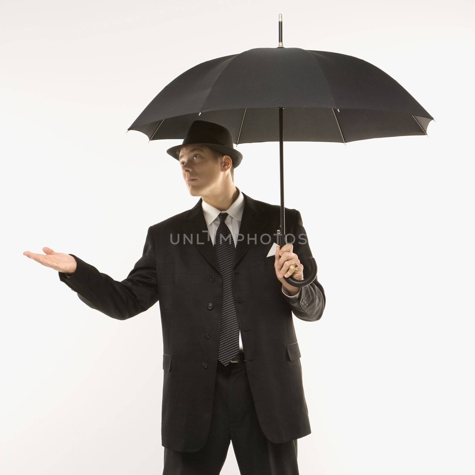 Caucasian mid-adult businessman wearing fedora holding umbrella with arm outstretched.