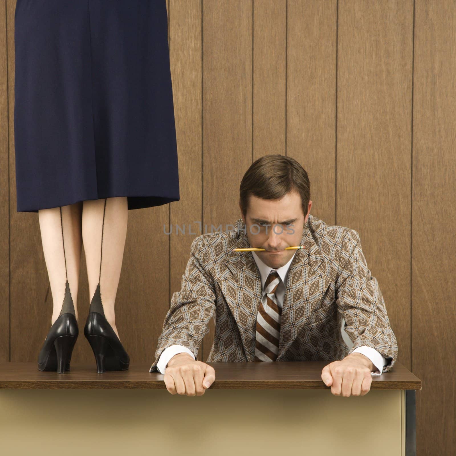 Mid-adult Caucasian male biting pencil and holding desk with Caucasian female standing on desk beside him.