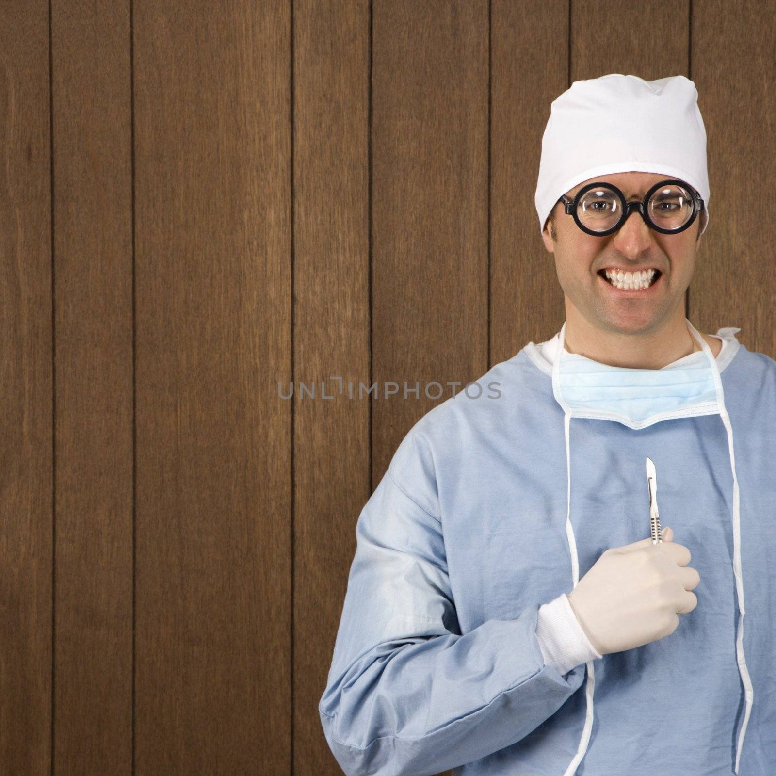 Mid-adult Caucasian male surgeon wearing thick glasses and holding scalpel smiling maniacally.