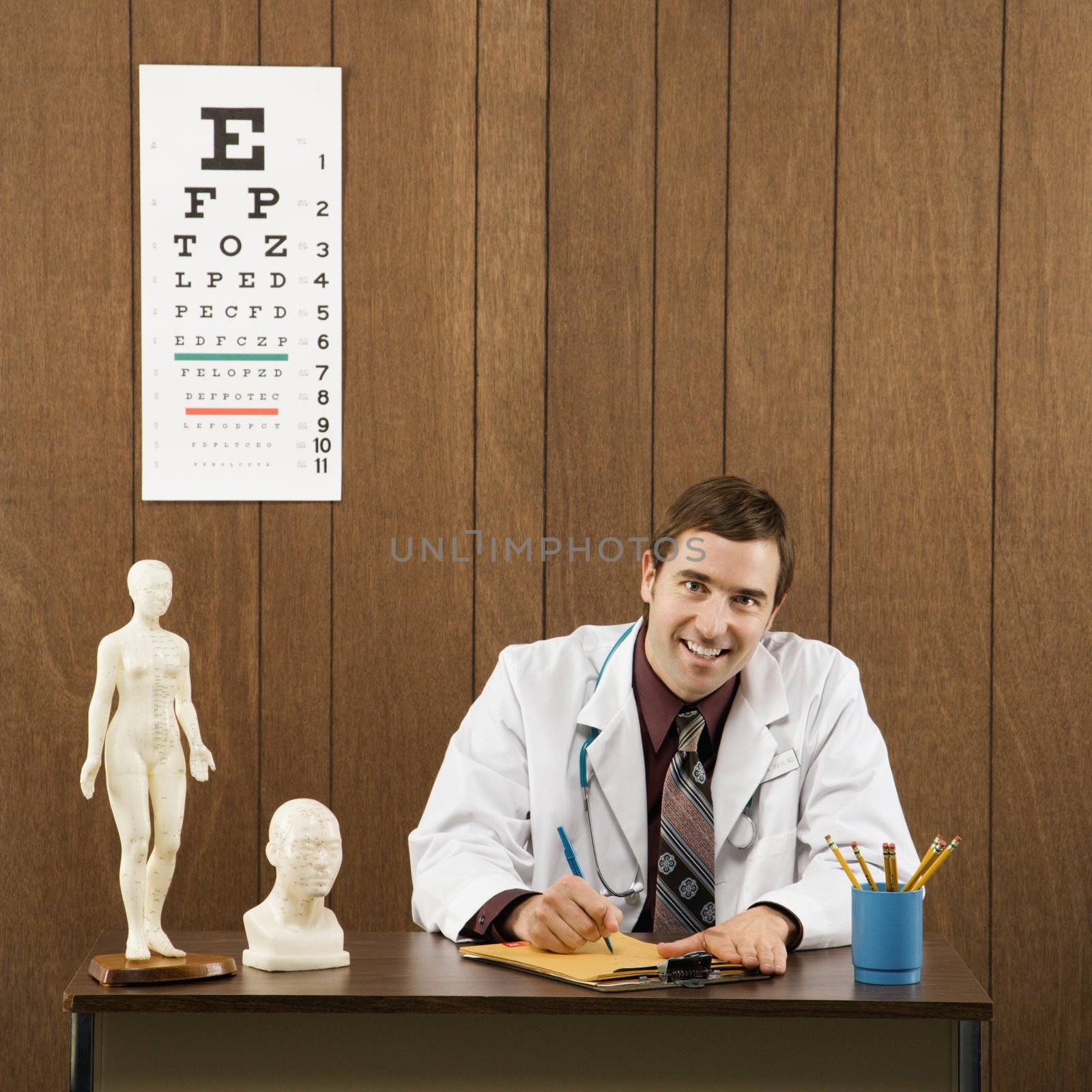 Mid-adult Caucasian male doctor sitting at desk writing and looking at viewer.