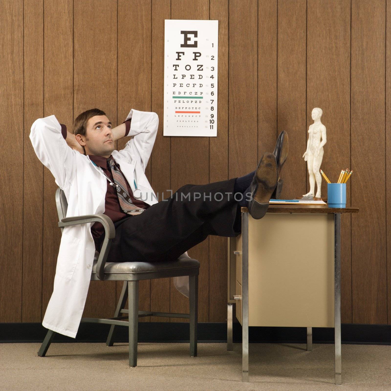 Mid-adult Caucasian male doctor at desk reclining with hands behind head and feet on desk.