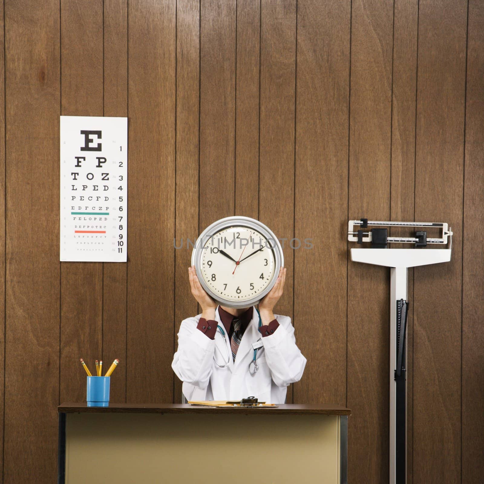 Caucasian male doctor sitting at desk holding clock over face.