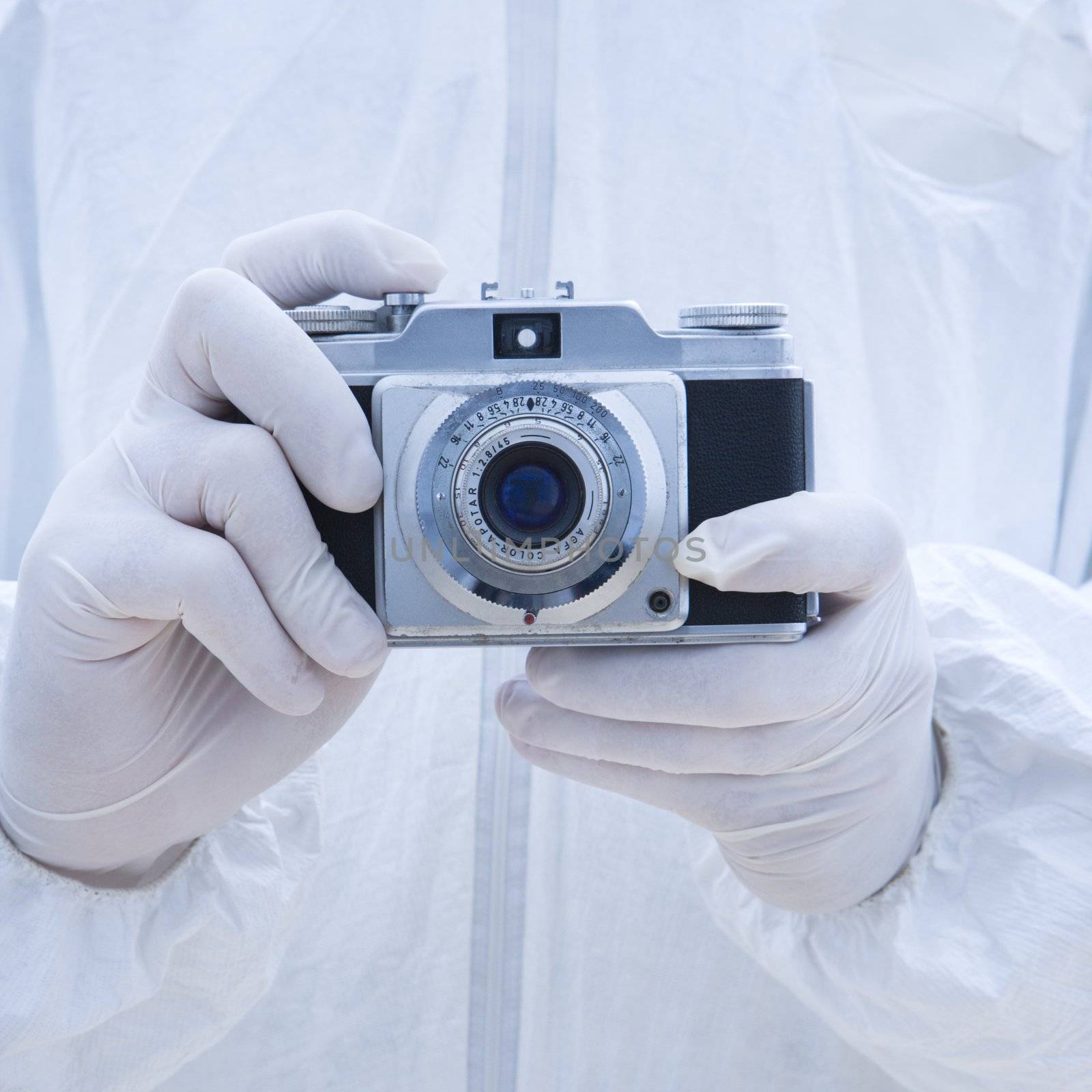 Man in biohazard suit holding antique camera with finger on shutter button.