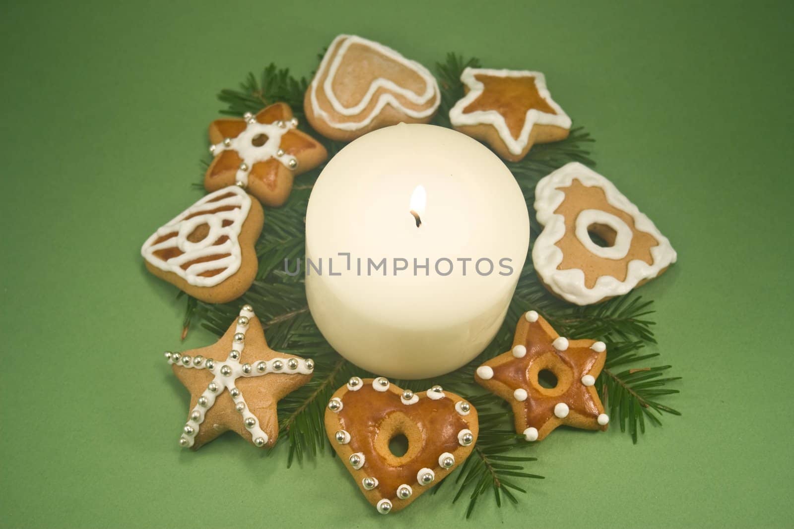 White pillar candle on fir branches surrounded by decorated gingerbread cookies on green background