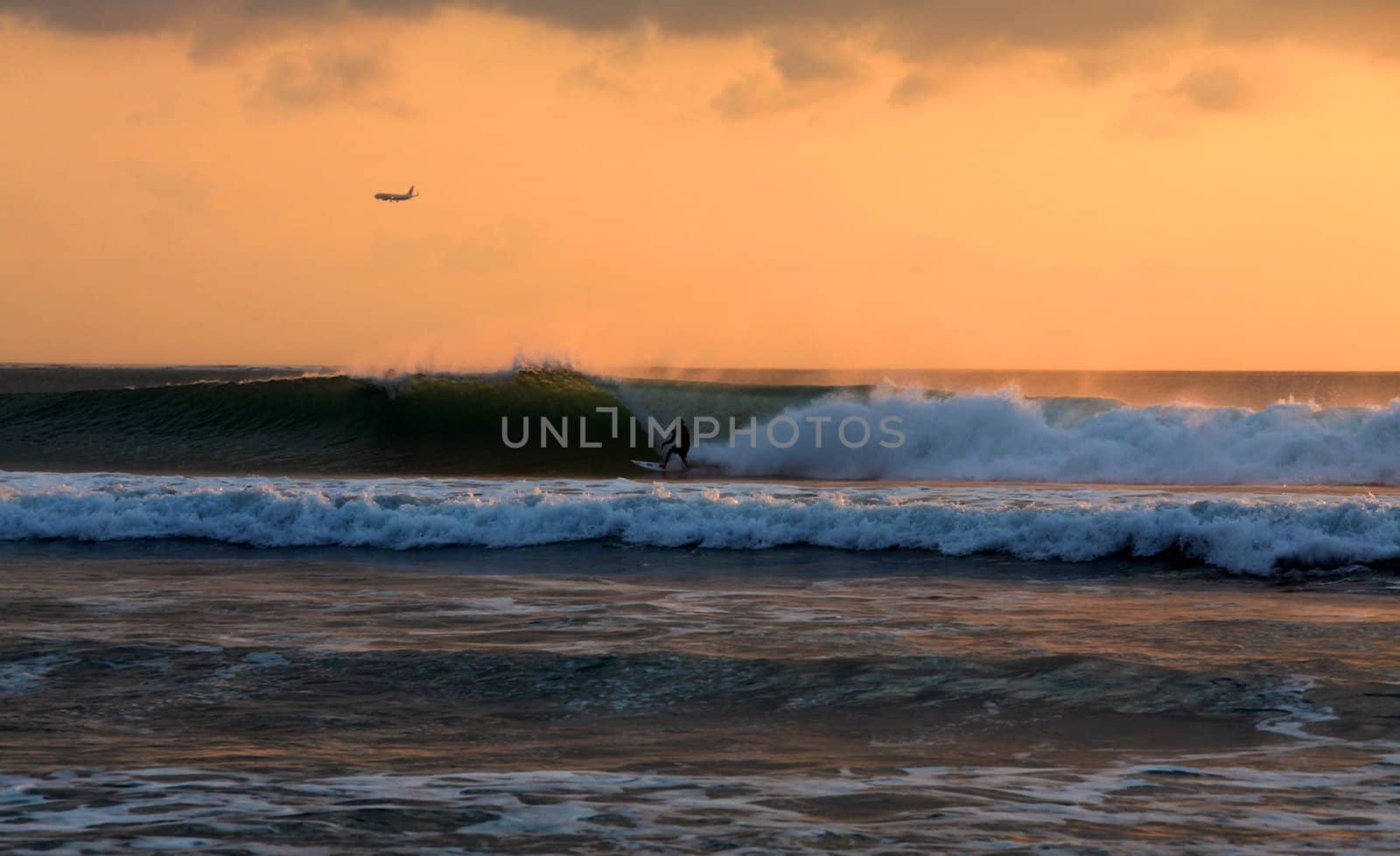 Surfer rides waves at sunset, airplane landing in the background