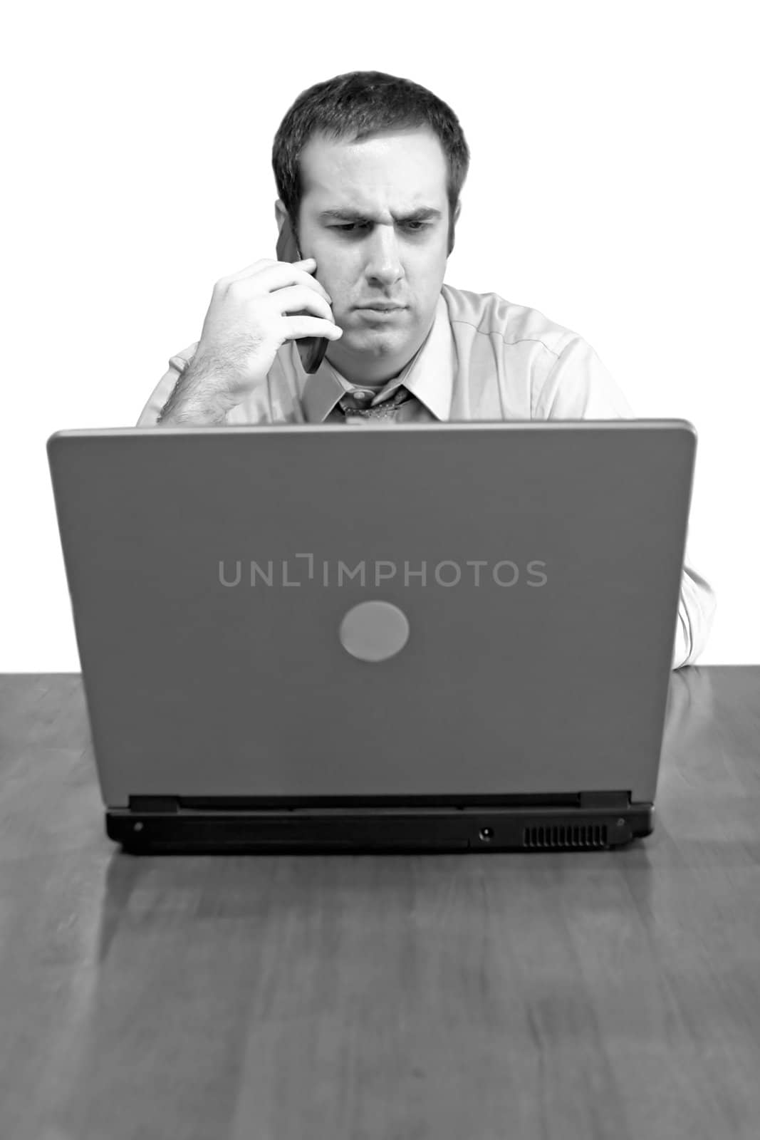 A man working from home with his cell phone and laptop in black and white. He has an upset or serious look on his face.