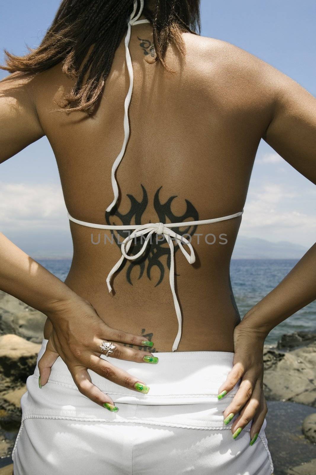 Rear view of multi ethnic young adult woman on beach with hands on hips and tattoo.