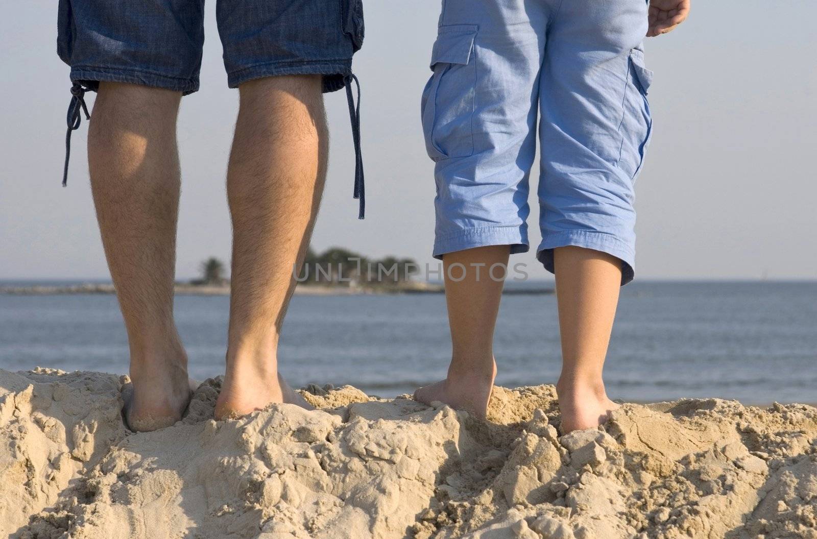 Legs of father and son at the top of a sand dune, walking towards the sea