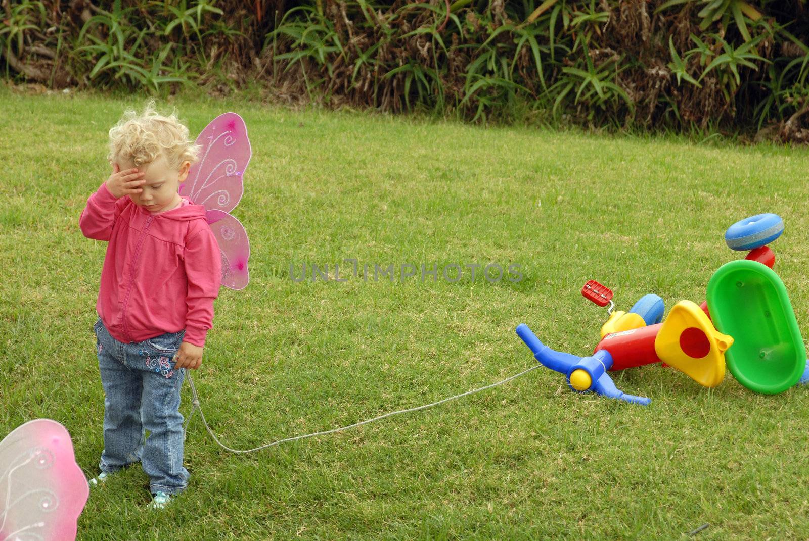 Blondie little girl with short curly hair on butterfly wings costume playing pretending sadness with tricycle on the grass in the park.