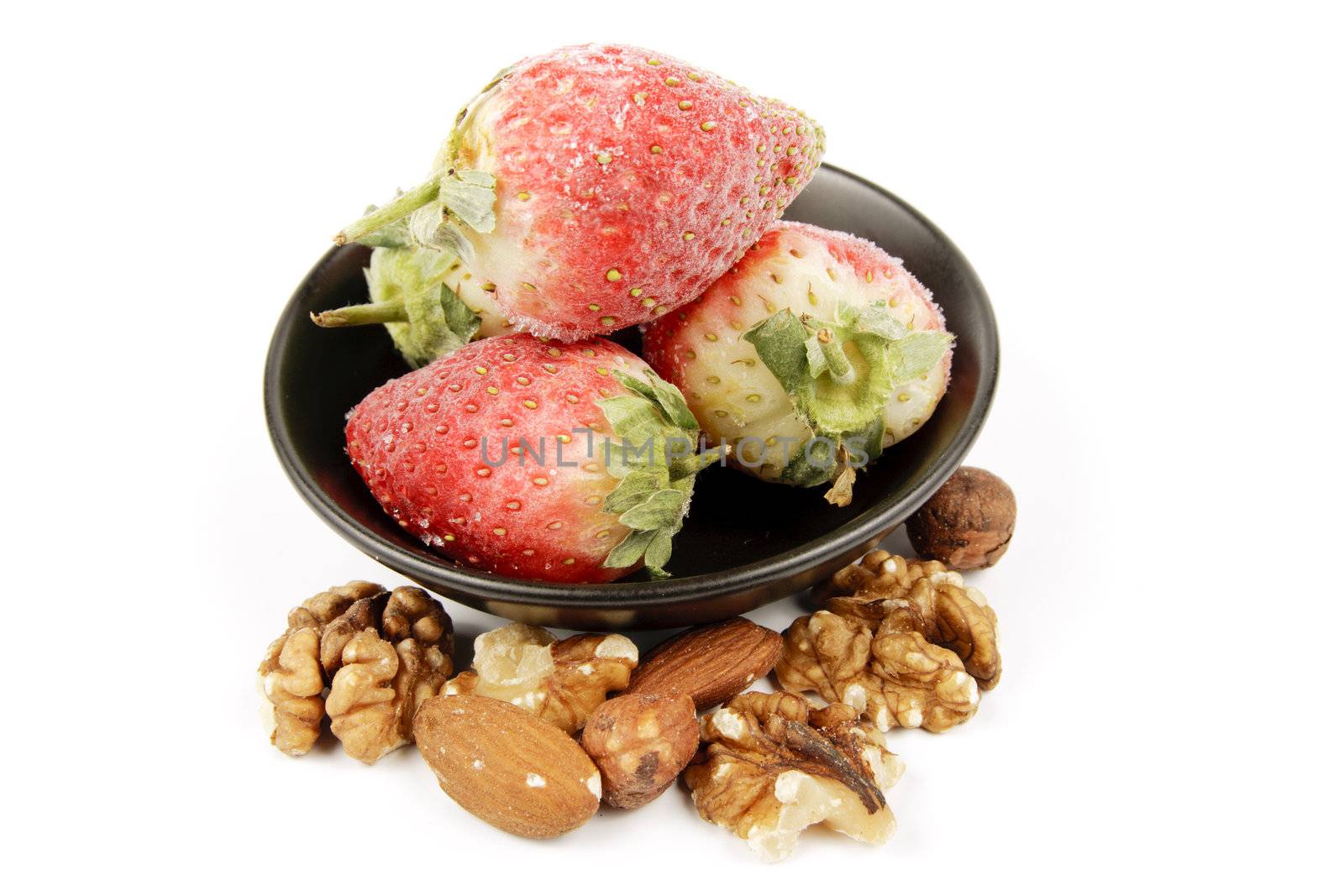 Red ripe frozen strawberries in a small black bowl with mixed nuts on a reflective white background