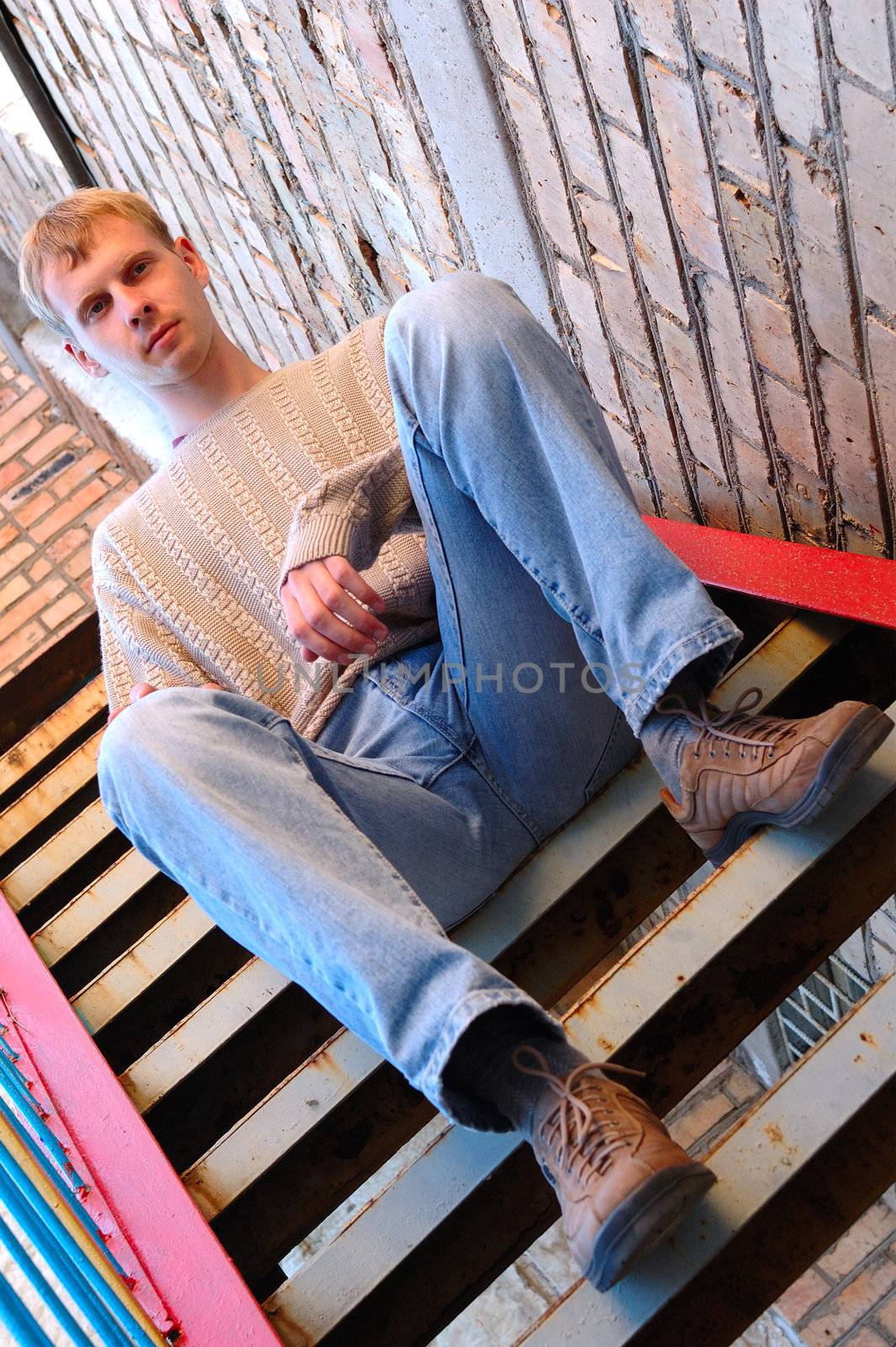Young stylish man with blonde hair sit on stairs near brick wall.