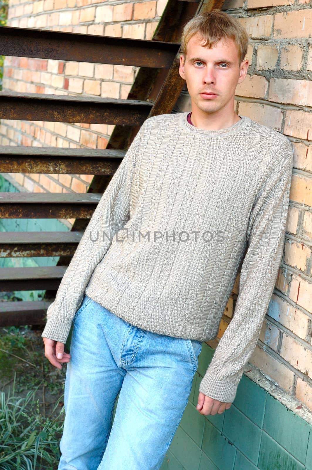 Young stylish man with blonde hair stay under stairs near brick wall.