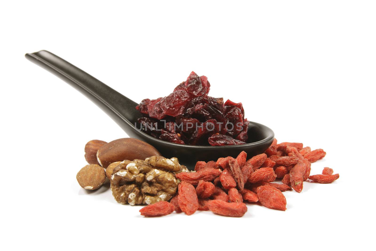 Red ripe dried cranberries on a small black spoon with mixed nuts and goji berries on a reflective white background