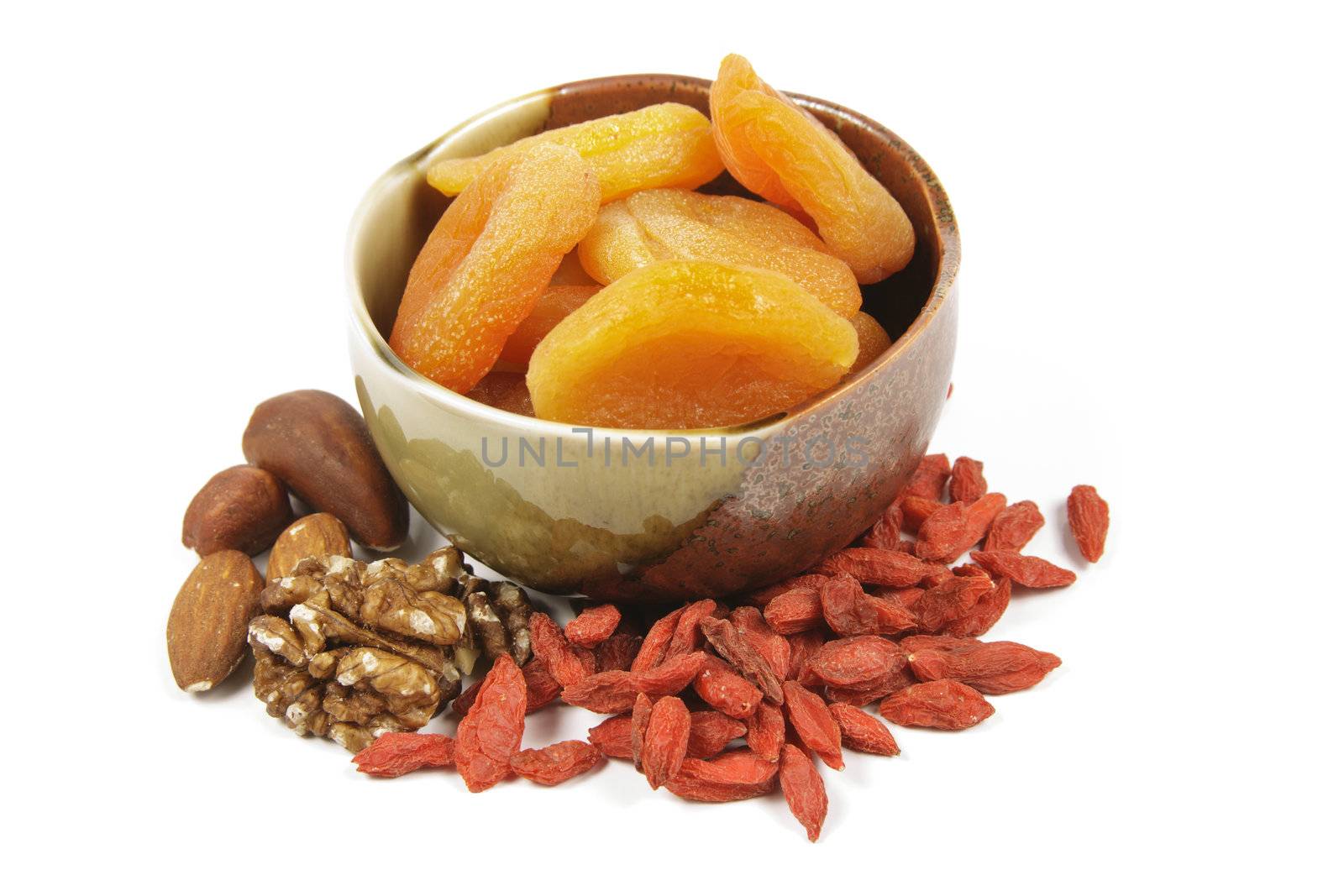 Dried juicy orange apricots in a green and brown bowl with mixed nuts and goji berries on a reflective white background