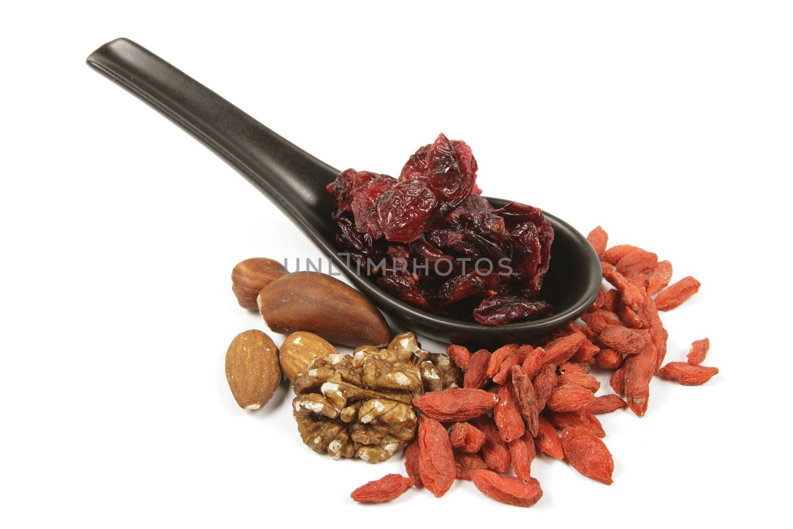 Red ripe dried cranberries on a small black spoon with mixed nuts and goji berries on a reflective white background