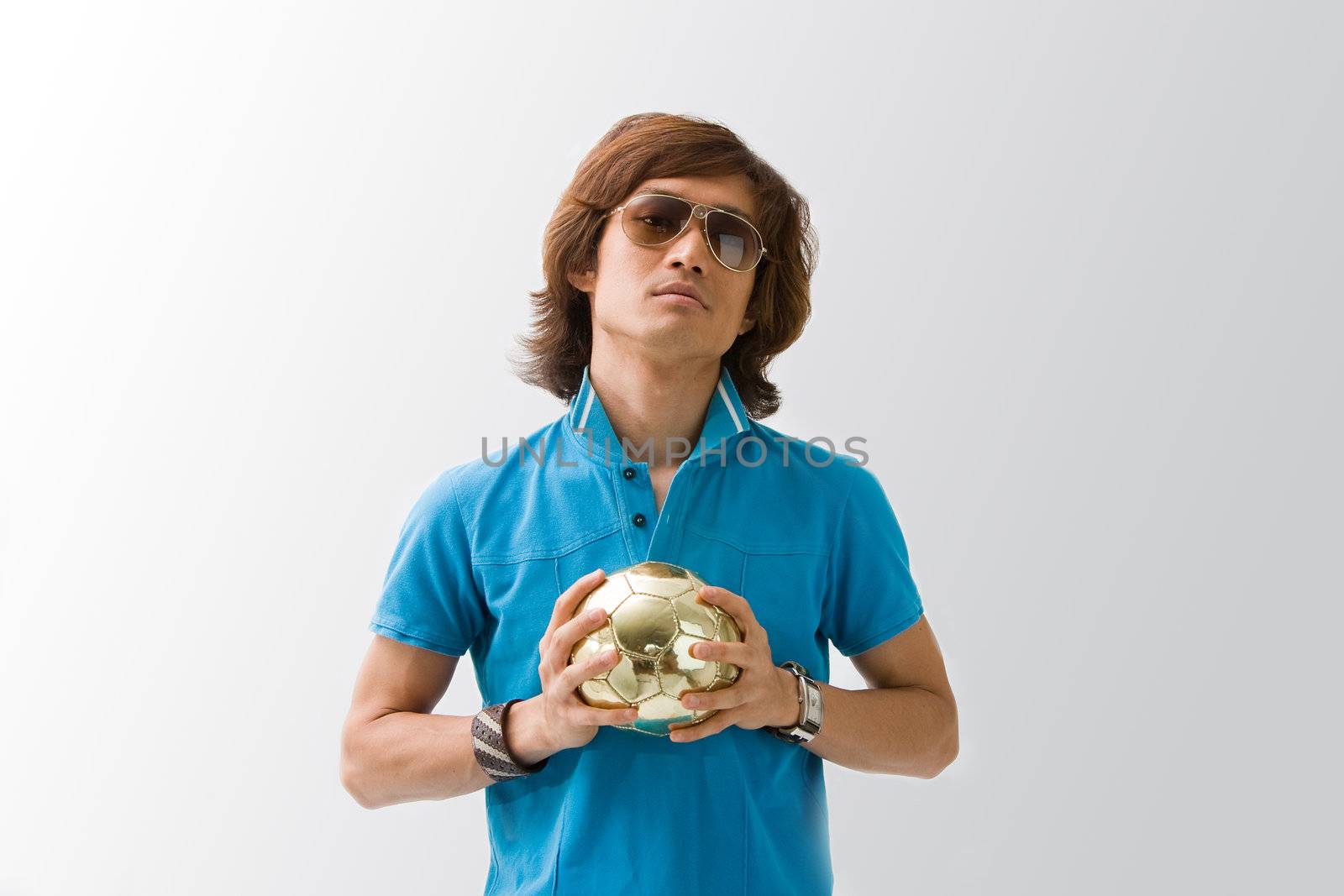 Sporty Asian guy in blue t-shirt holding a small golden soccer ball with both hands, isolated