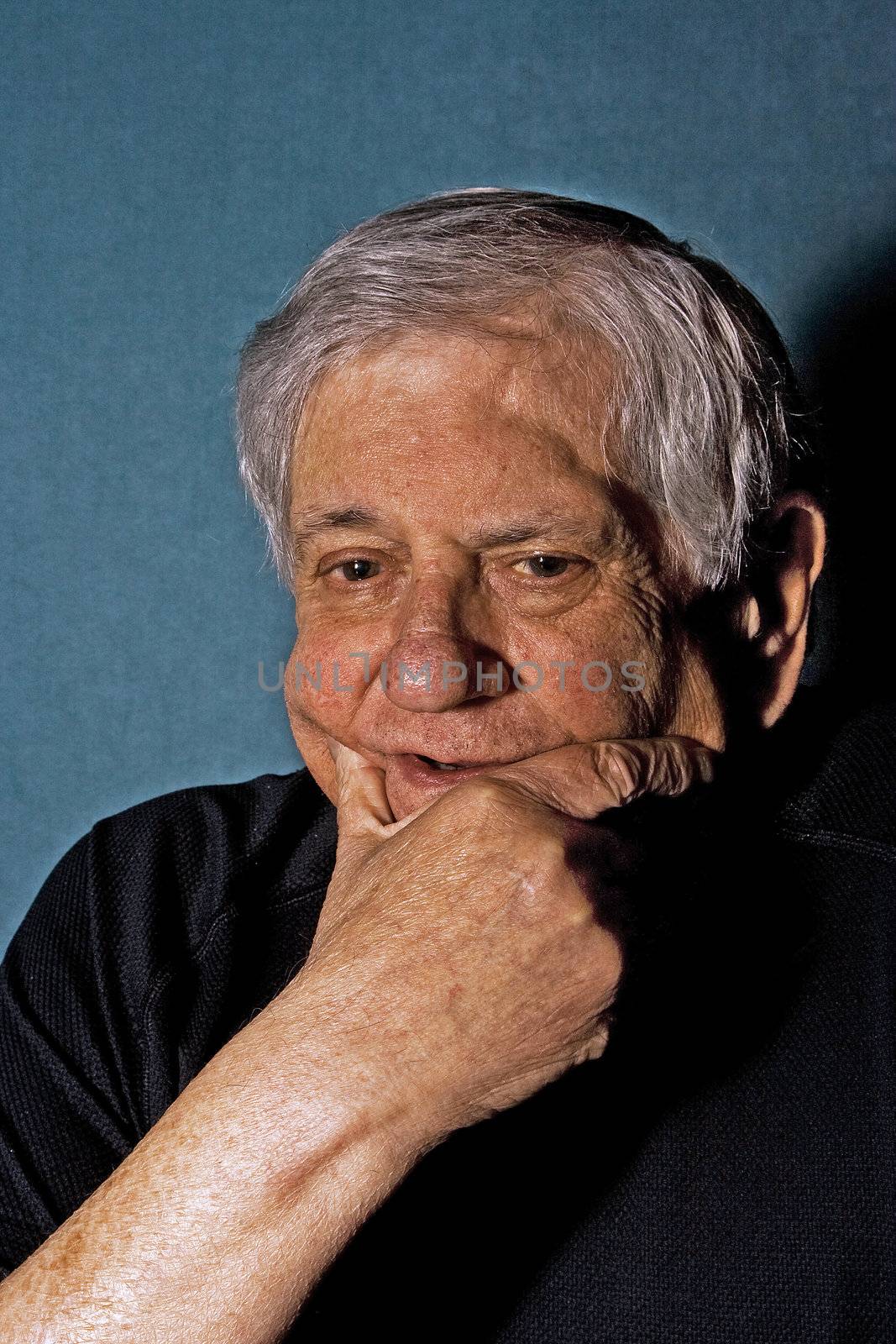 Dramatic portrait of a senior man with his hand on the side of his face wearing a black shirt isolated on gray/blue
