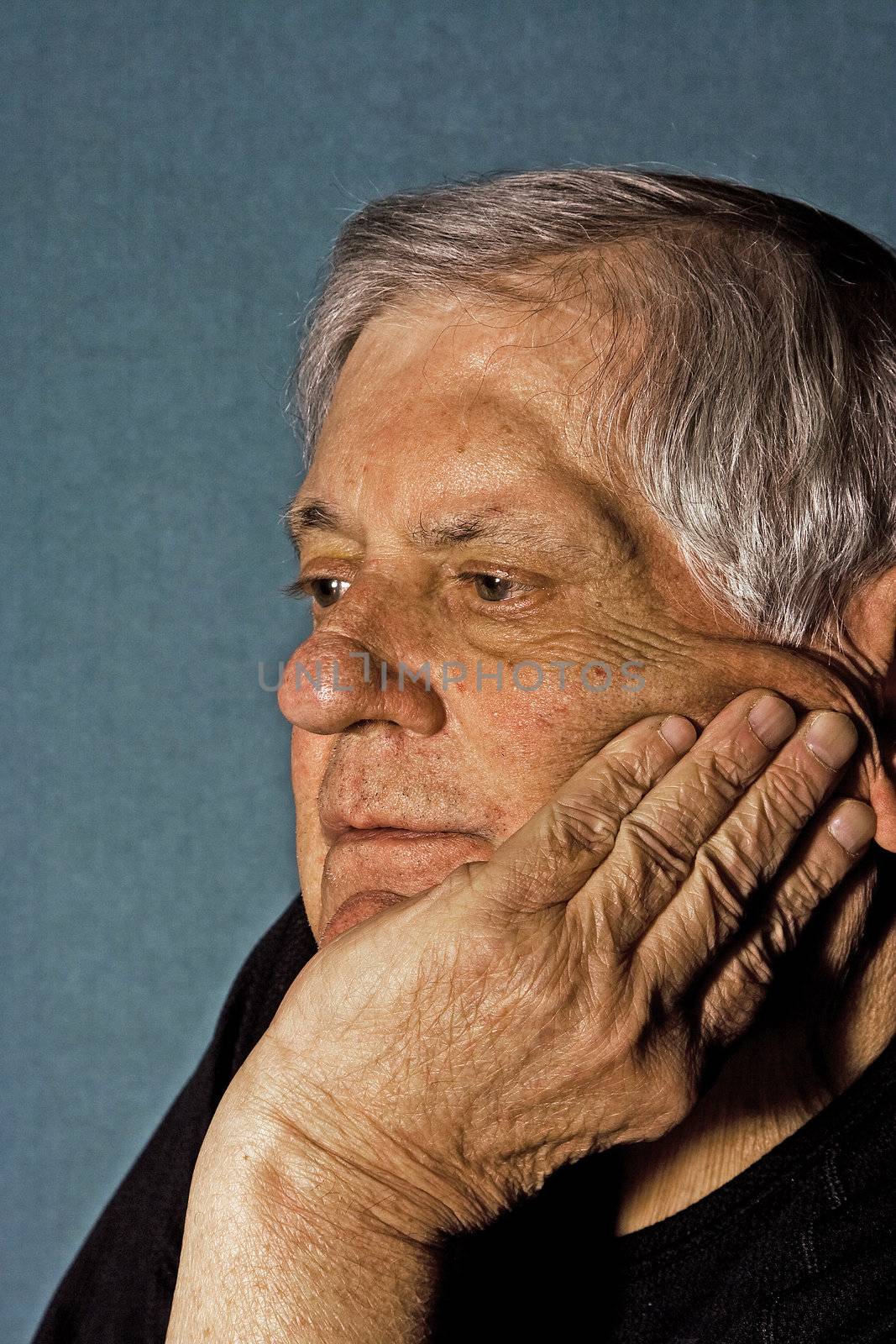 Dramatic side portrait of a senior man with his hand on the side of his face wearing a black shirt isolated on gray/blue