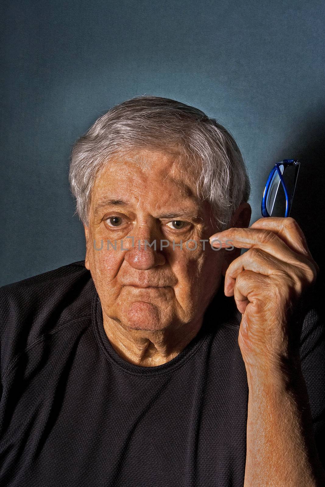 Dramatic side portrait of a senior man with a pair of glasses in his hand next to his face wearing a black shirt isolated on gray/blue