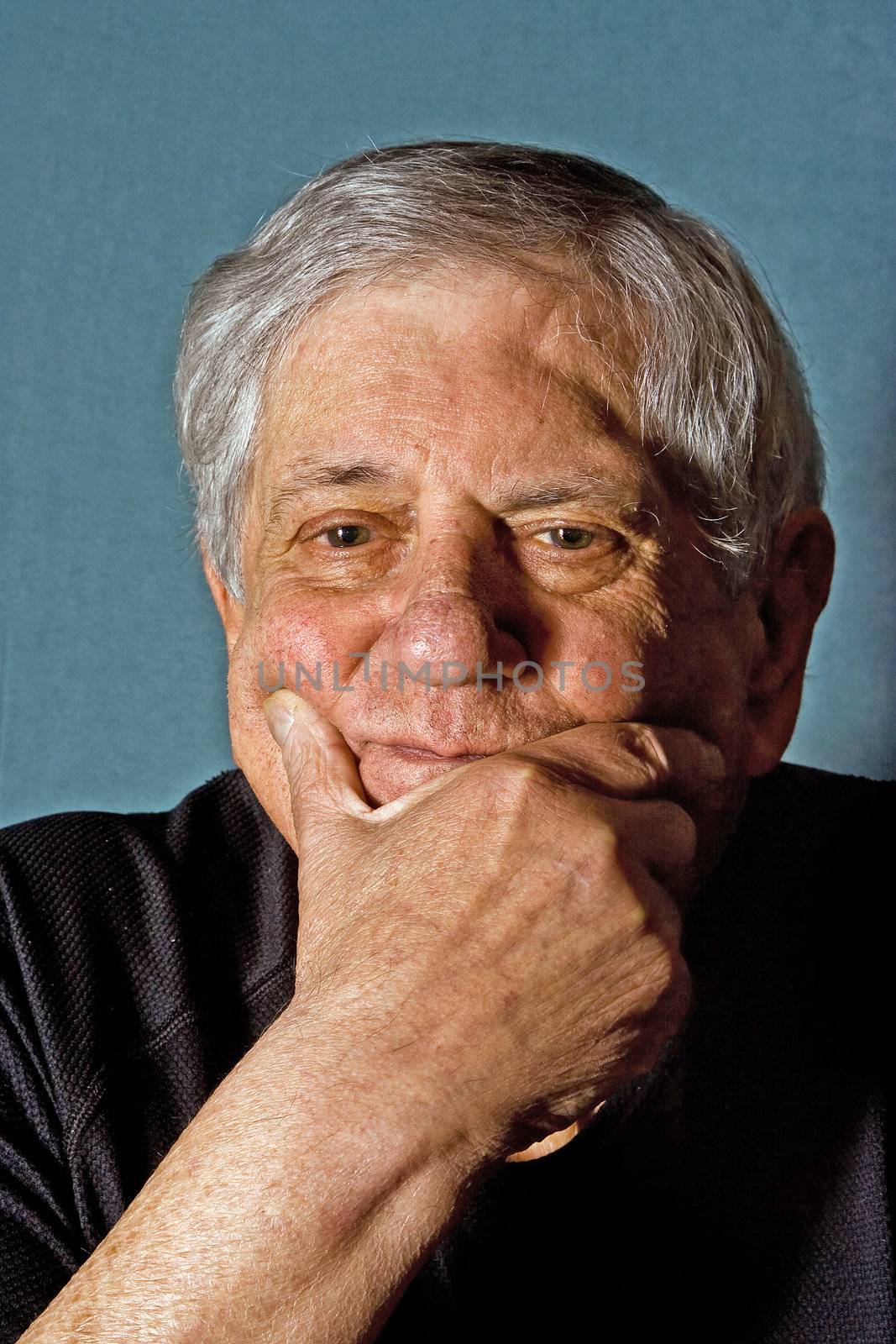 Dramatic portrait of a senior man with his hand on his chin wearing a black shirt isolated on gray/blue