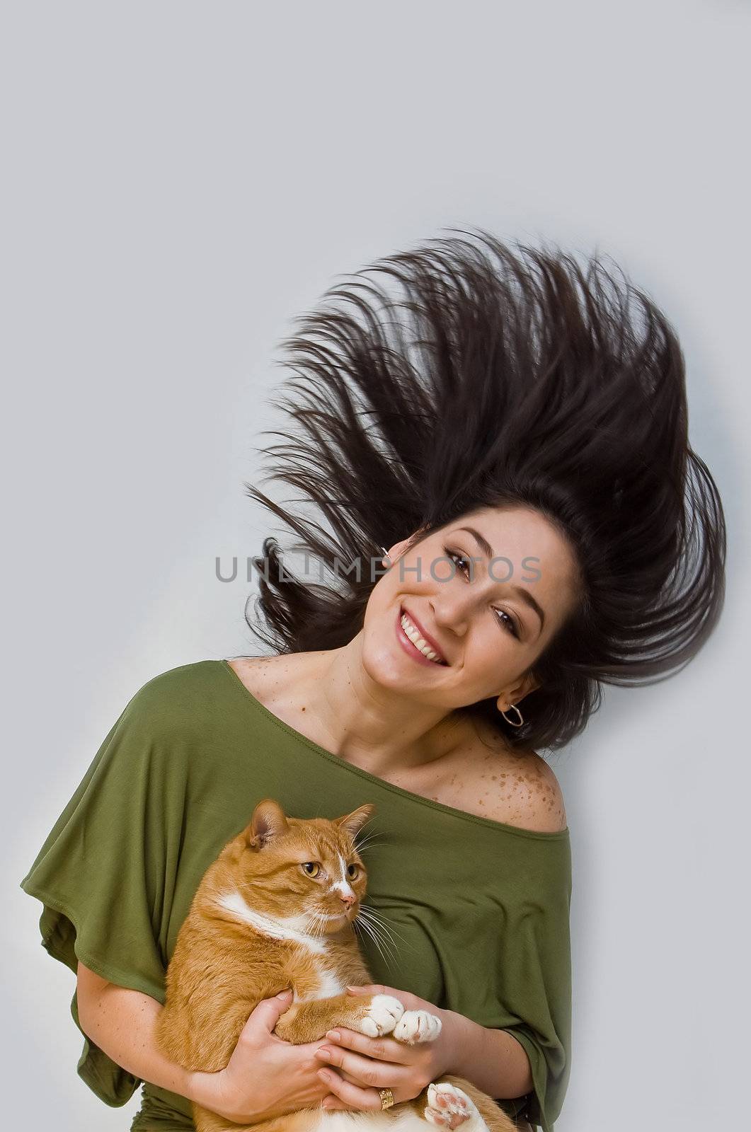 A woman with big smile in green shirt swinging her dark brown hair around while holding an orange cat, isolated on white