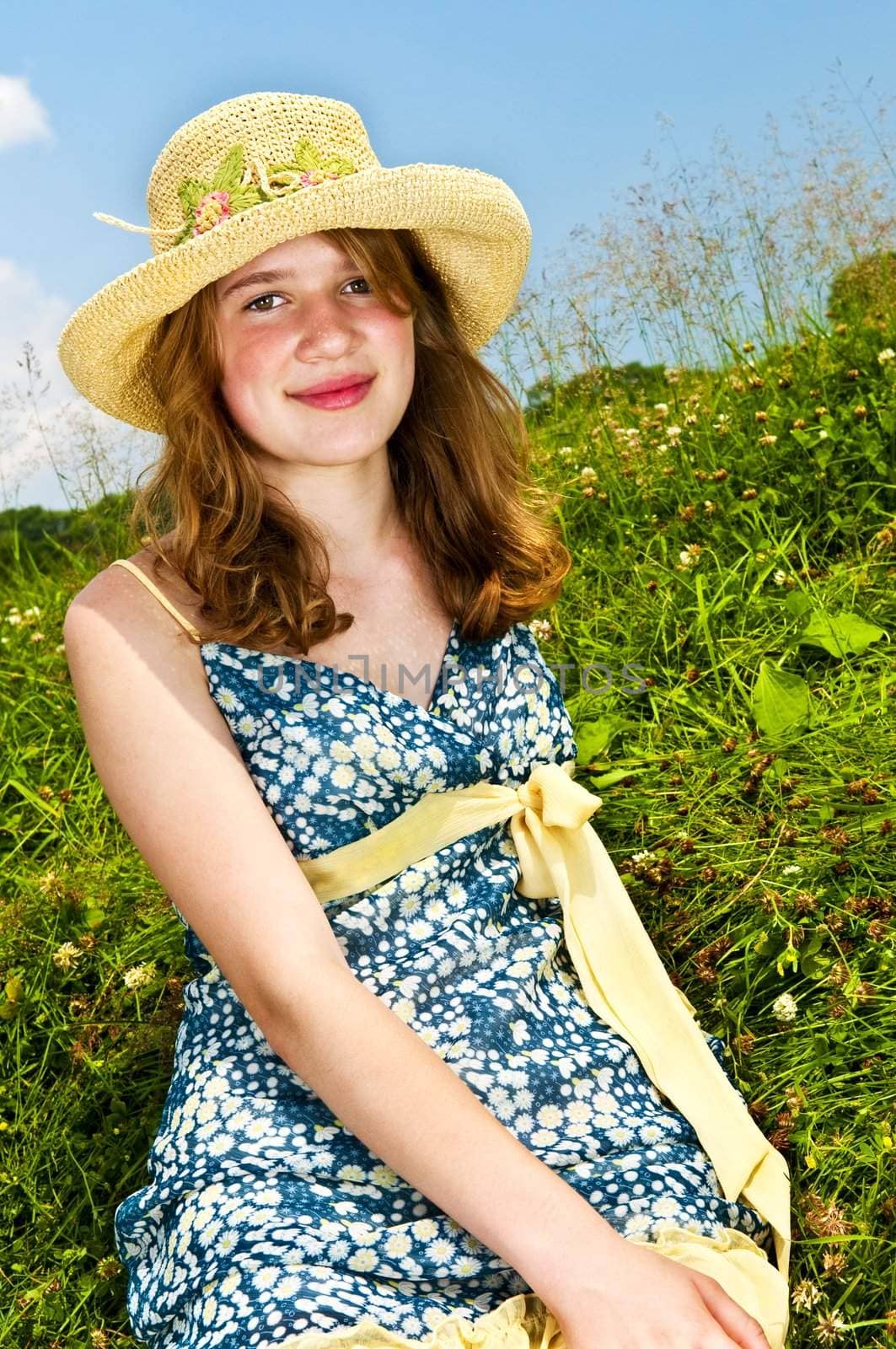 Young teenage girl sitting on summer meadow in straw hat