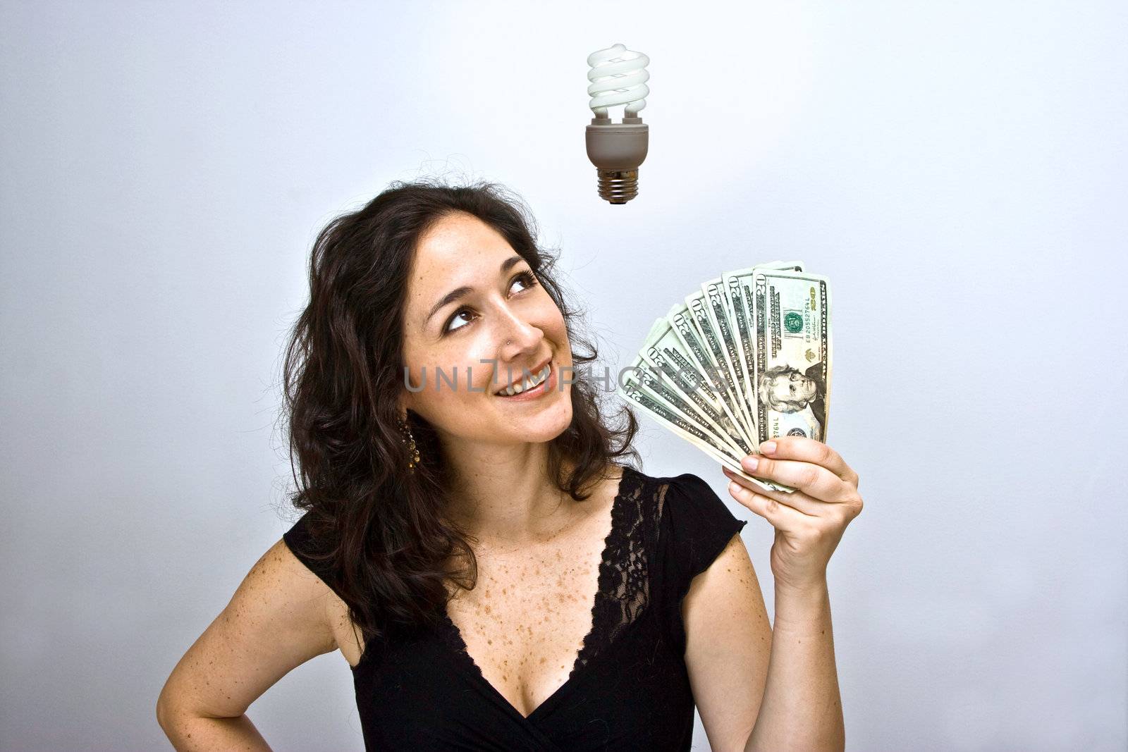 Woman waving money and looking up. Having an environmentally friendly idea with an energy saving fluorescent light bulb floating above her head, on a white background