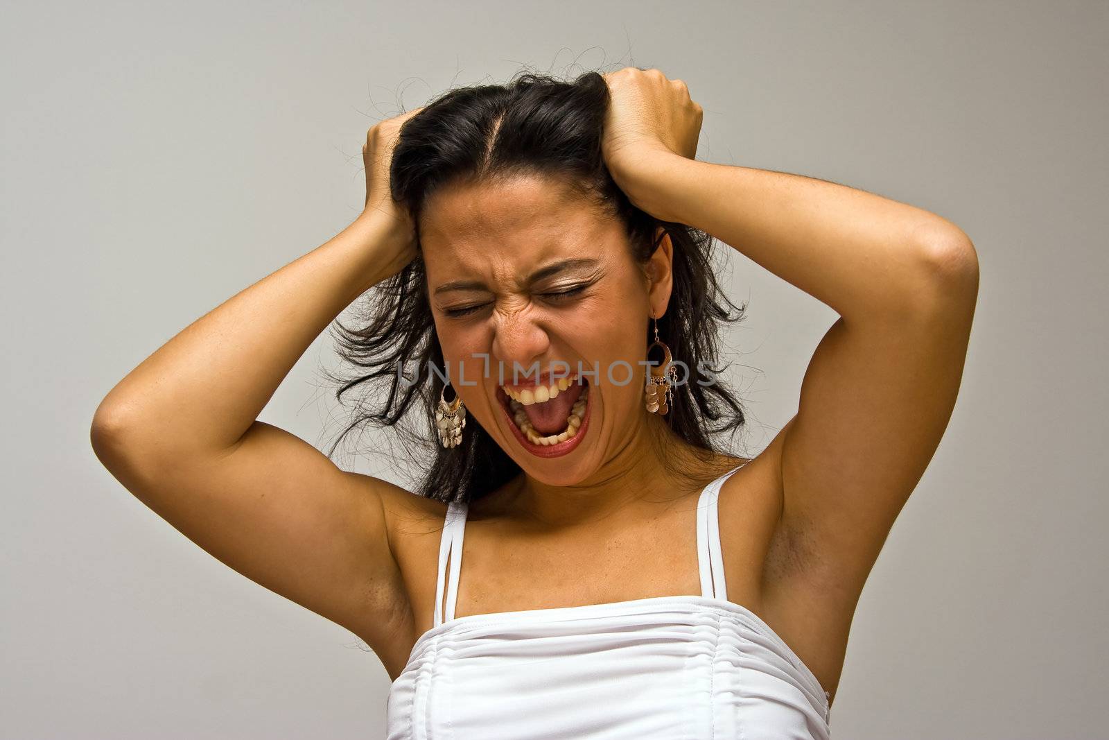 A young beautiful latina woman screeming of frustration and pulling her hair with both hands wearing a white shirt, isolated on white