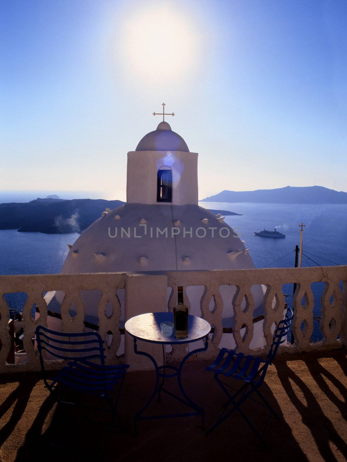 A balcony overlooking a small church on the Greek island of Santorini,  with the sun setting directly in front blue sky and mountains in the background.