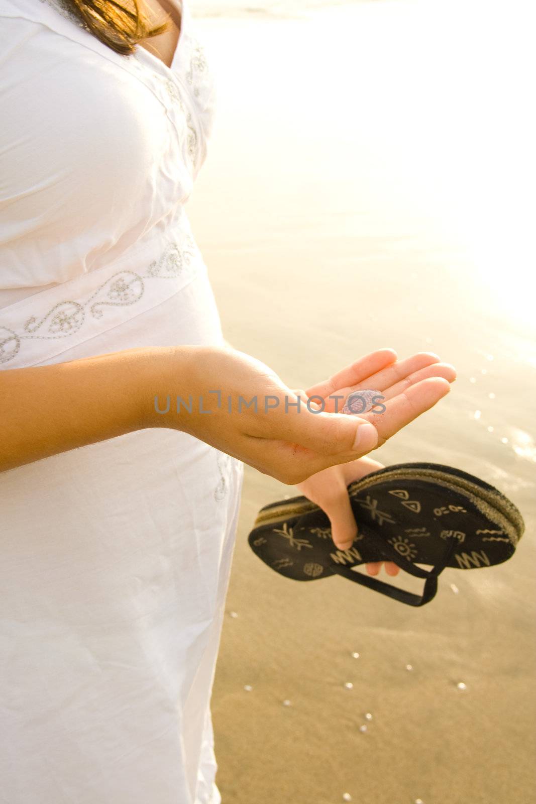 Pregnant woman standing on beach, holding shell and sandals.