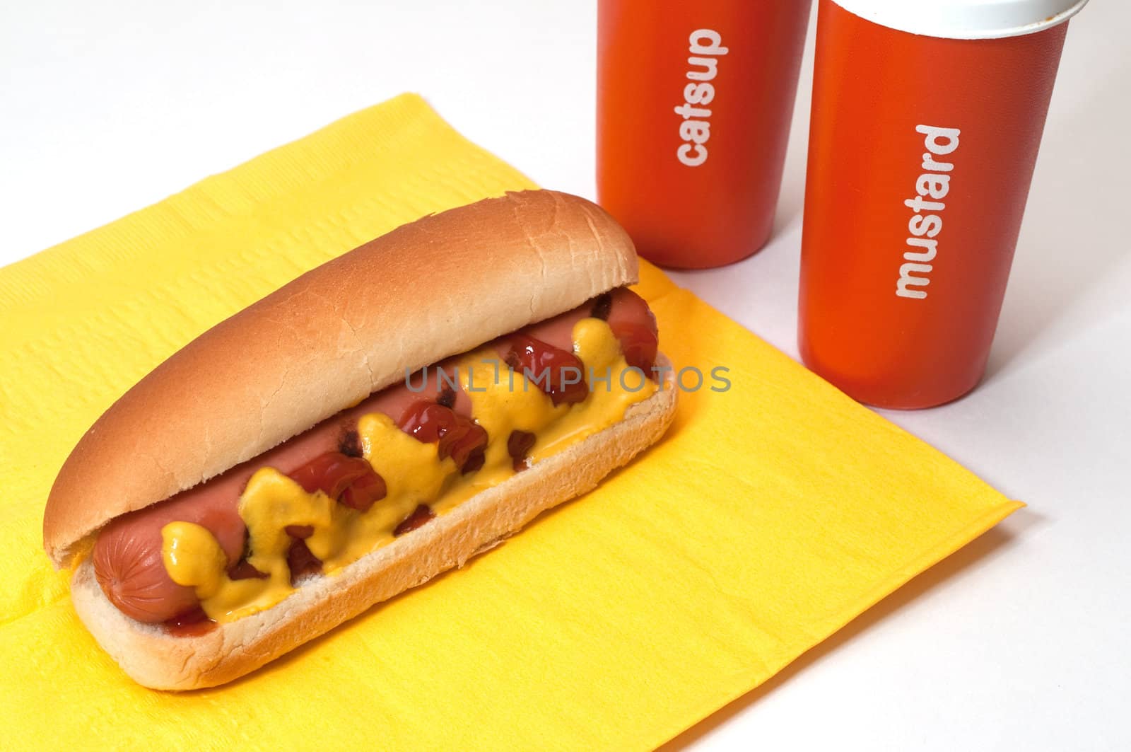 Hot dog with ketchup and mustard on yellow napkin.  Condiment bottles in background. 