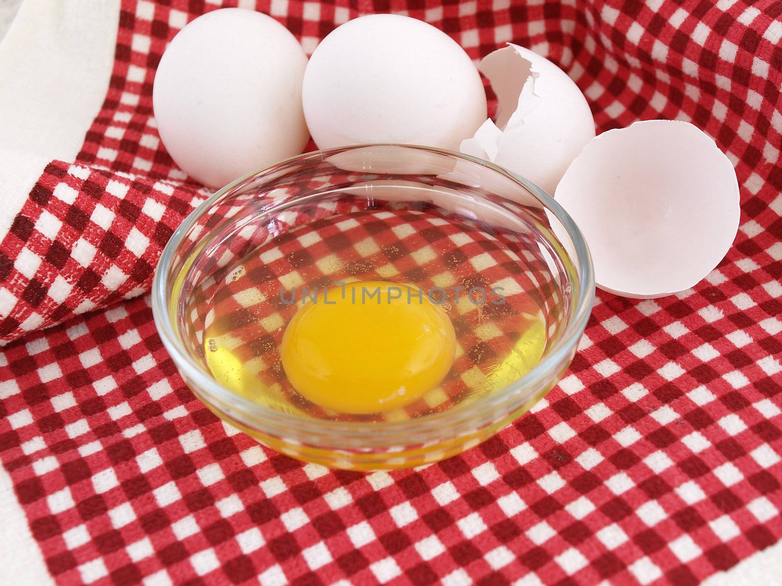 An egg cracked into a dish, eggshells and whole eggs laying on a red checkered background.