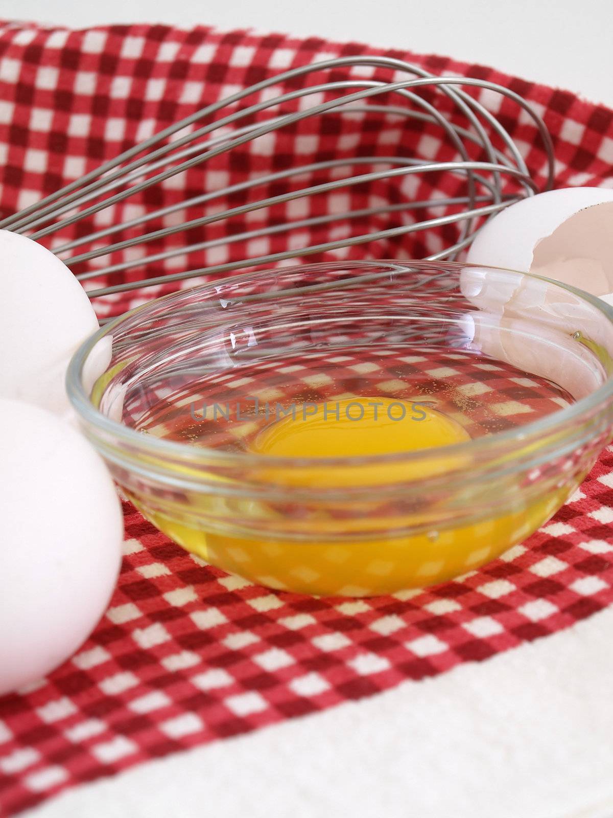 An egg cracked into a dish, eggshells and whole eggs with a whisk laying on a red checkered background.