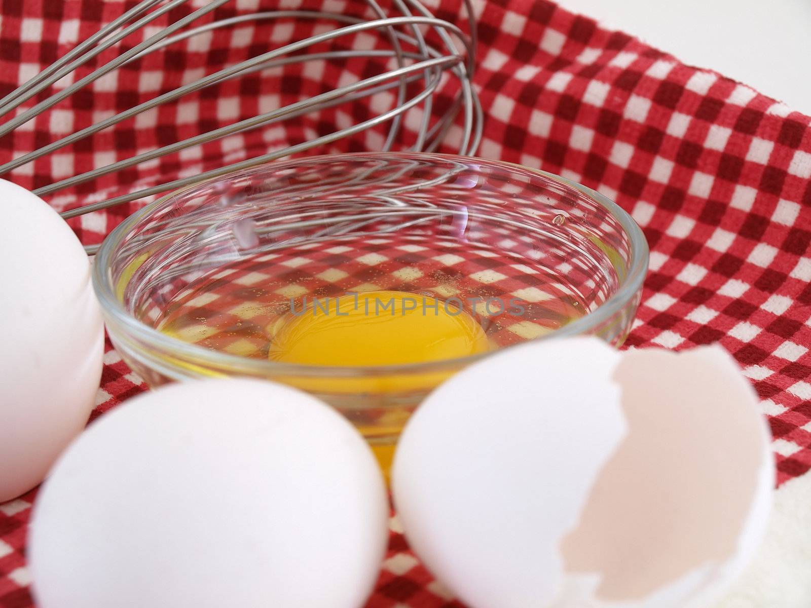 An egg cracked into a dish, eggshells and whole eggs with a whisk laying on a red checkered background.