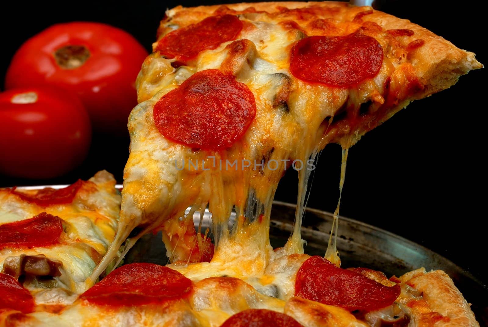 Slice of pepperoni pizza being removed from whole pizza with tomatoes in background.  Isolated on black background.