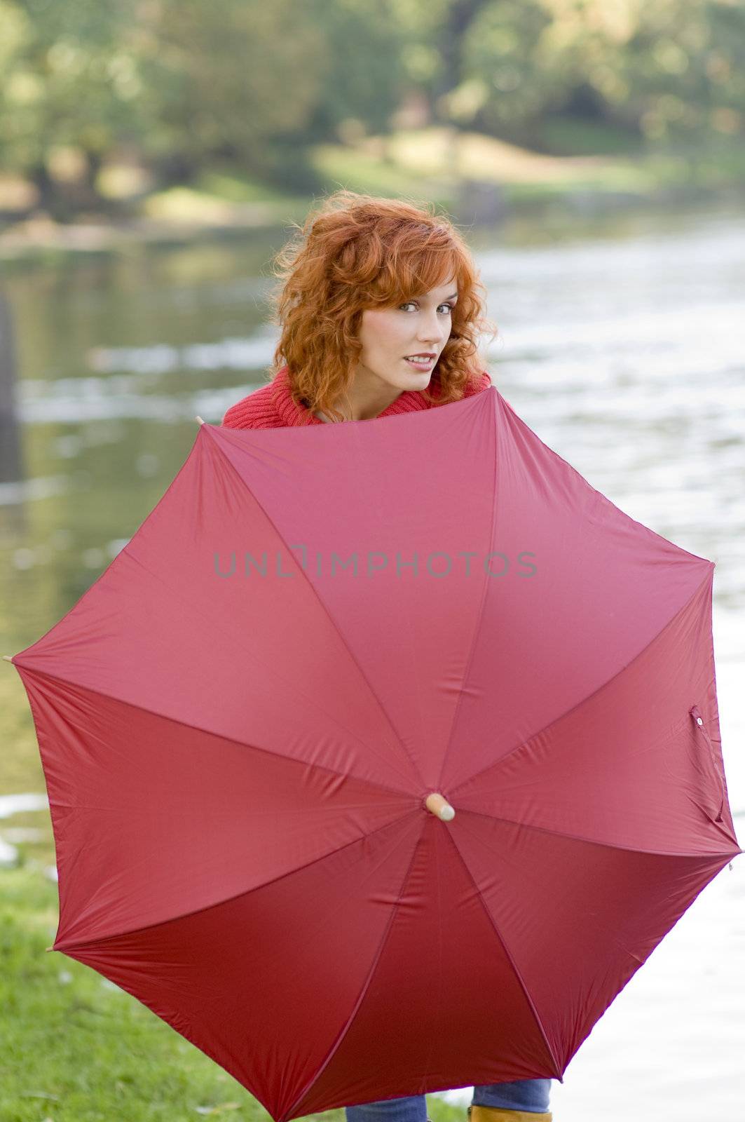 Pretty red haired girl near river playing behind a red umbrella