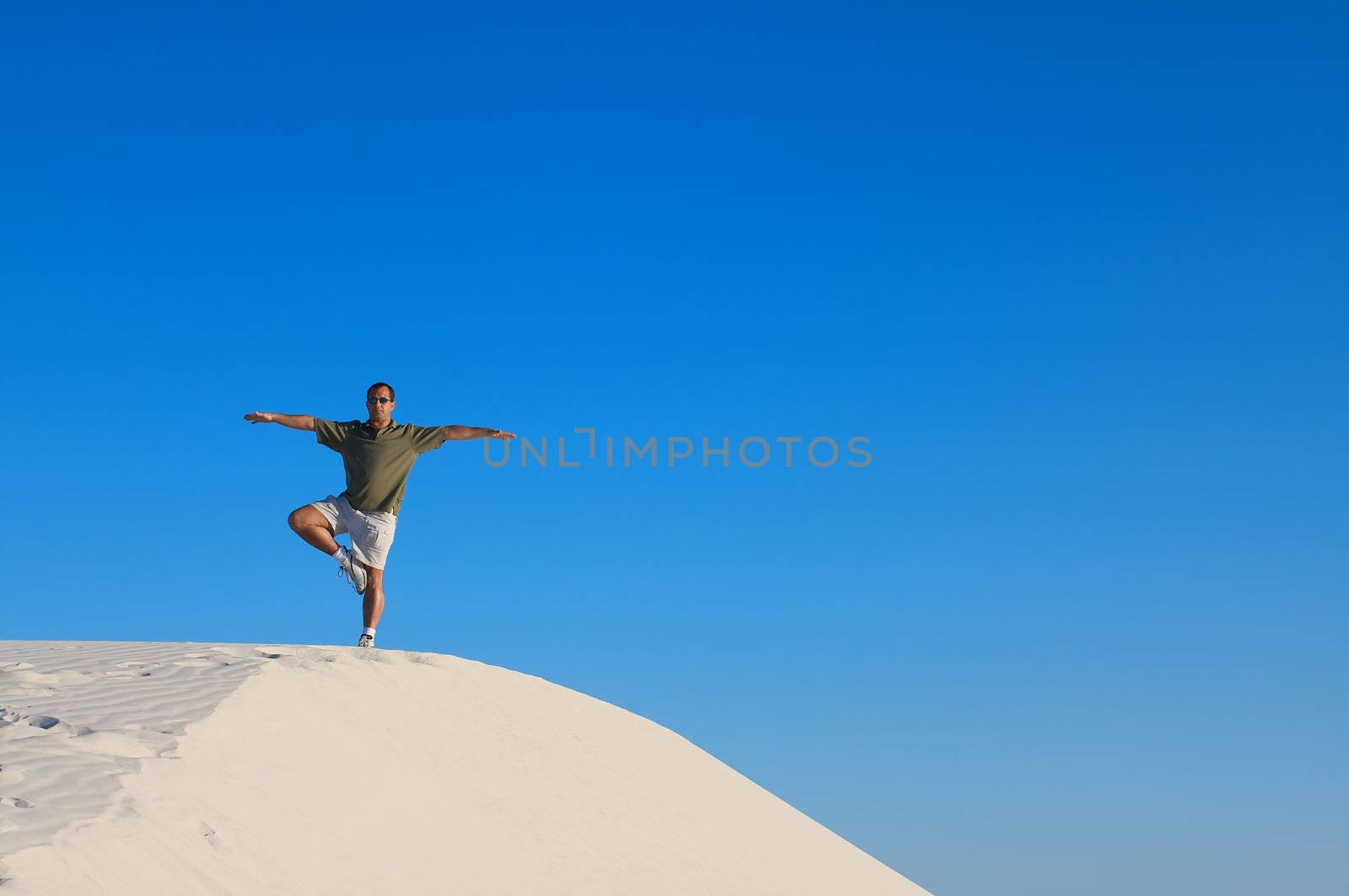 Man in a red shirt with his arms raised in a yoga tree pose looking off into the distance on top of a white sand dune with a blue sky