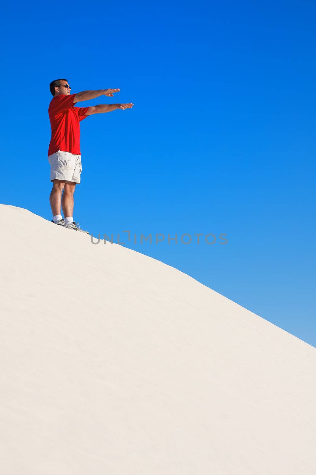 Man in a red shirt with his arms raised in a yoga pose looking off into the distance on top of a white sand dune with a blue sky