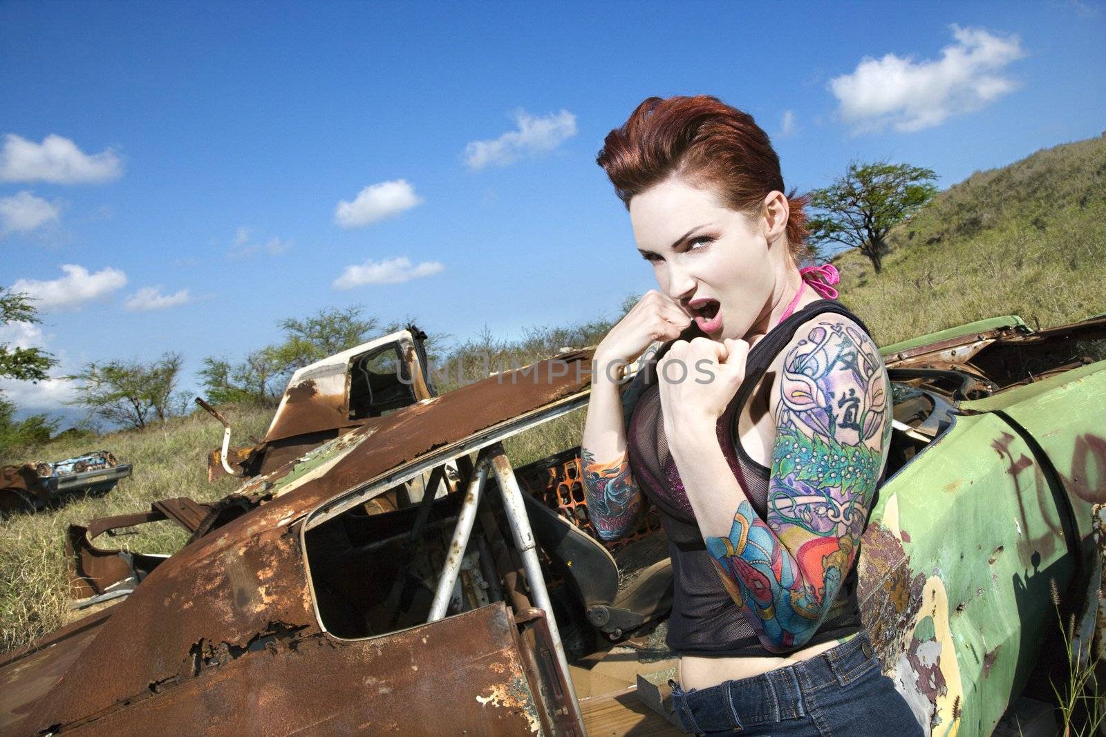 Angry tattooed Caucasian woman with fists clenched ready to fight in junkyard.